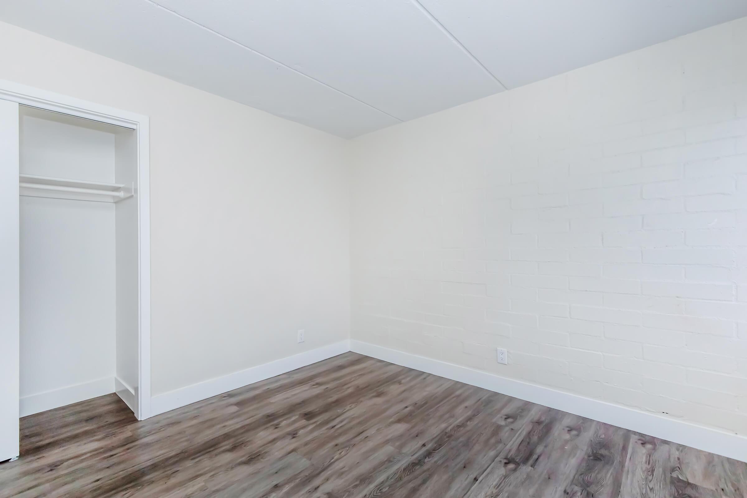 A vacant room with light-colored walls and a wooden floor. There is a white closet to the left with an open door, and the room appears bright and spacious, lacking any furniture or decorations.