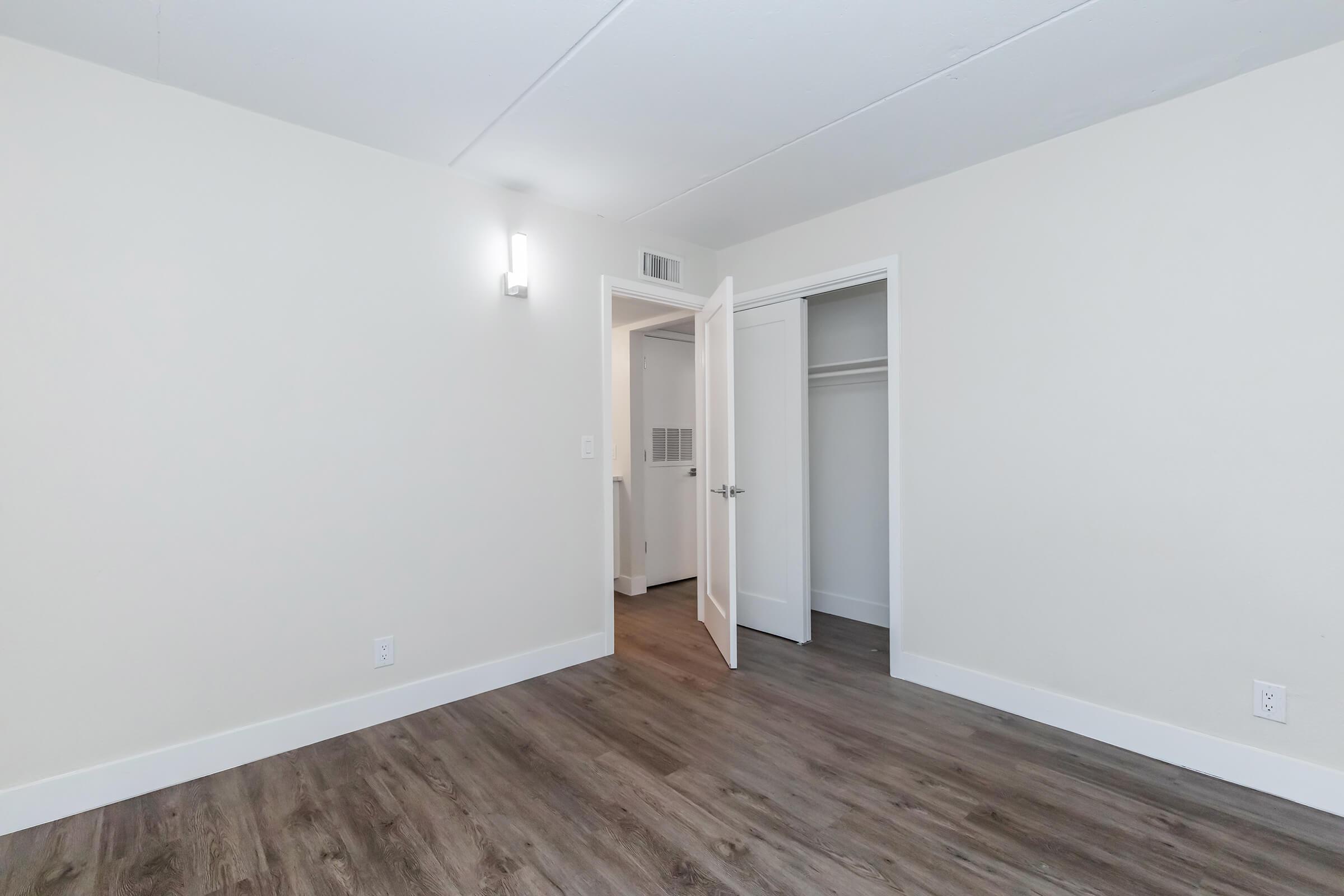 A clean, empty room featuring light-colored walls and a light wooden floor. There's an open white closet door on the left, and the room is brightly lit by a wall-mounted light fixture. The space has a simple, minimalist design with no furniture.