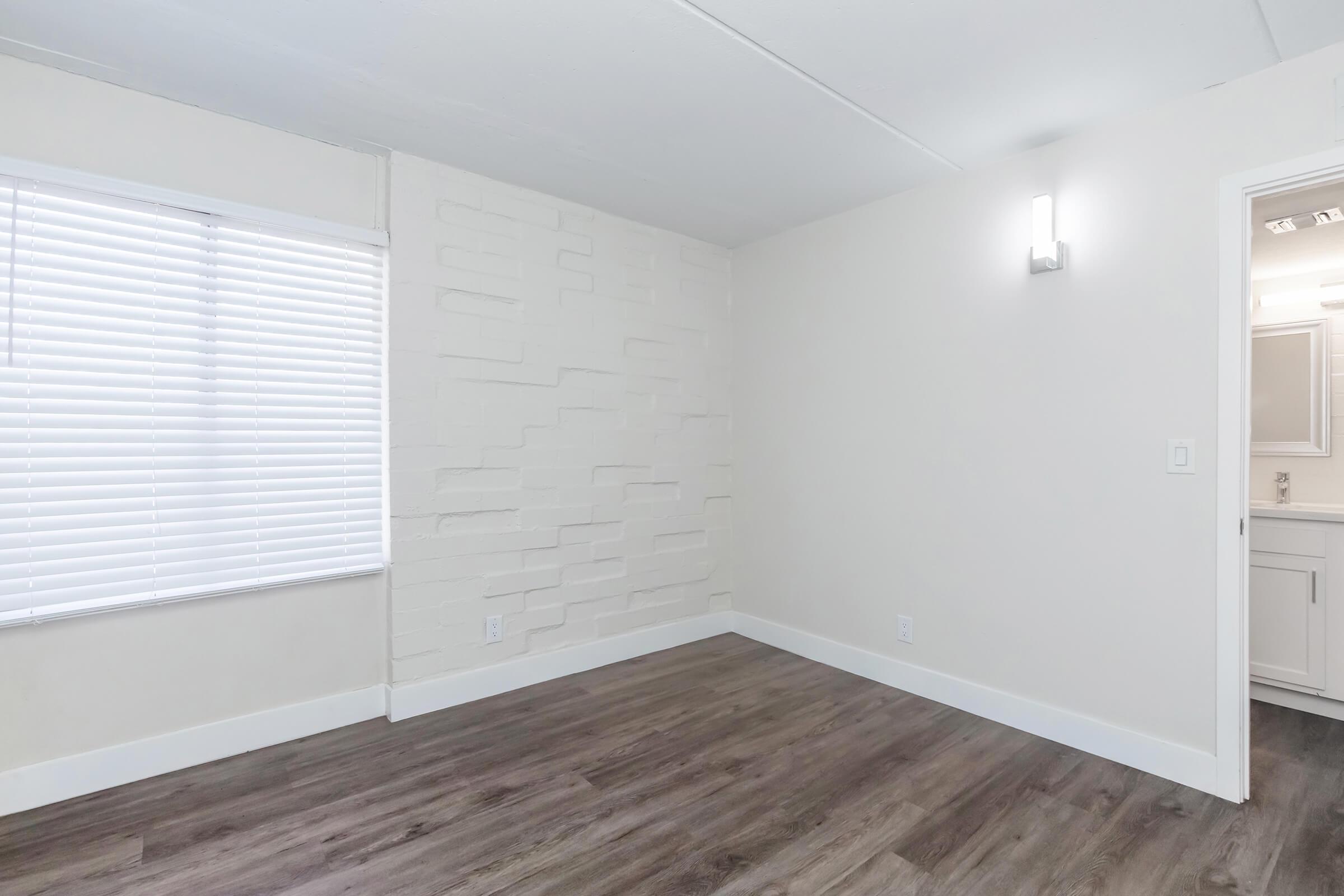 A minimalist interior room with a light-colored wall featuring a textured design. There is a window with horizontal blinds allowing natural light, and a light fixture mounted on the wall. The floor is made of light wood laminate, creating a clean and modern look. A doorway leads to a bathroom area visible in the background.