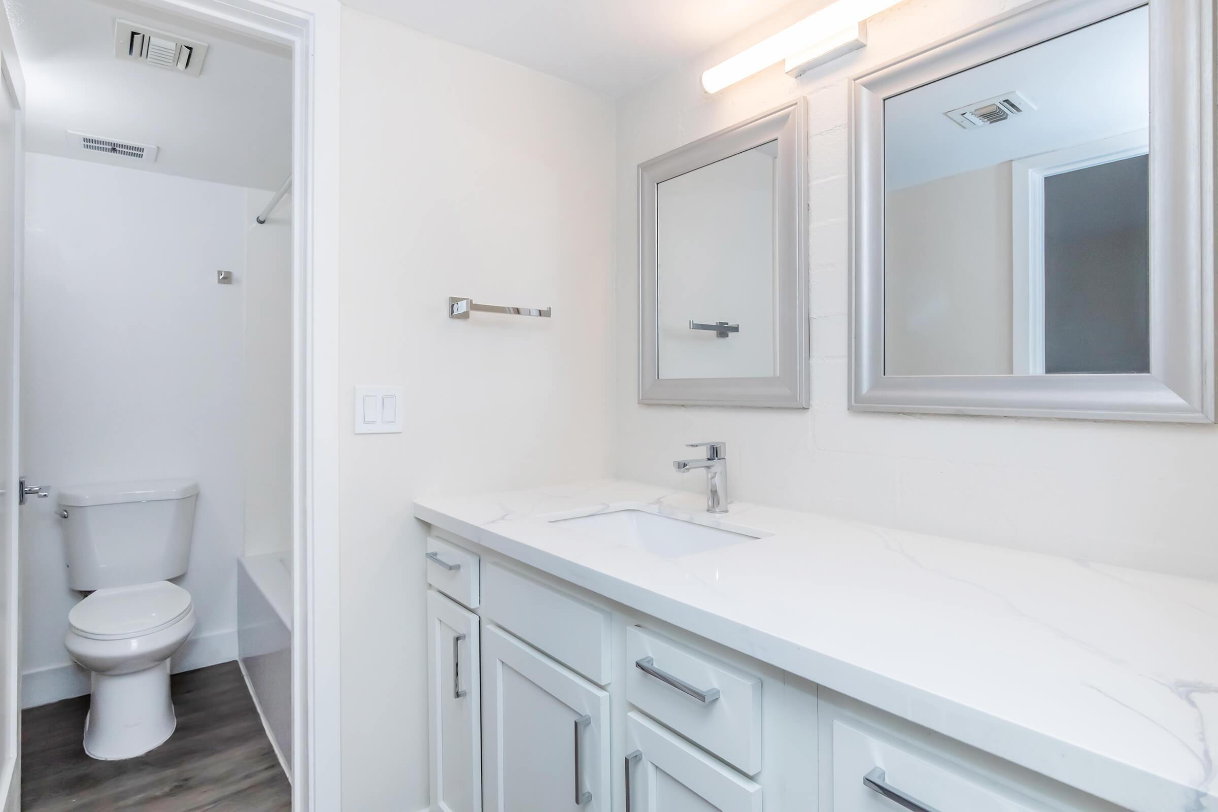 Modern bathroom featuring a double sink vanity with a white countertop, two mirrors, and light fixtures. A clean, minimalist design with light-colored walls and gray flooring. In the background, a separate area contains a toilet and shower space, offering a functional and stylish layout.