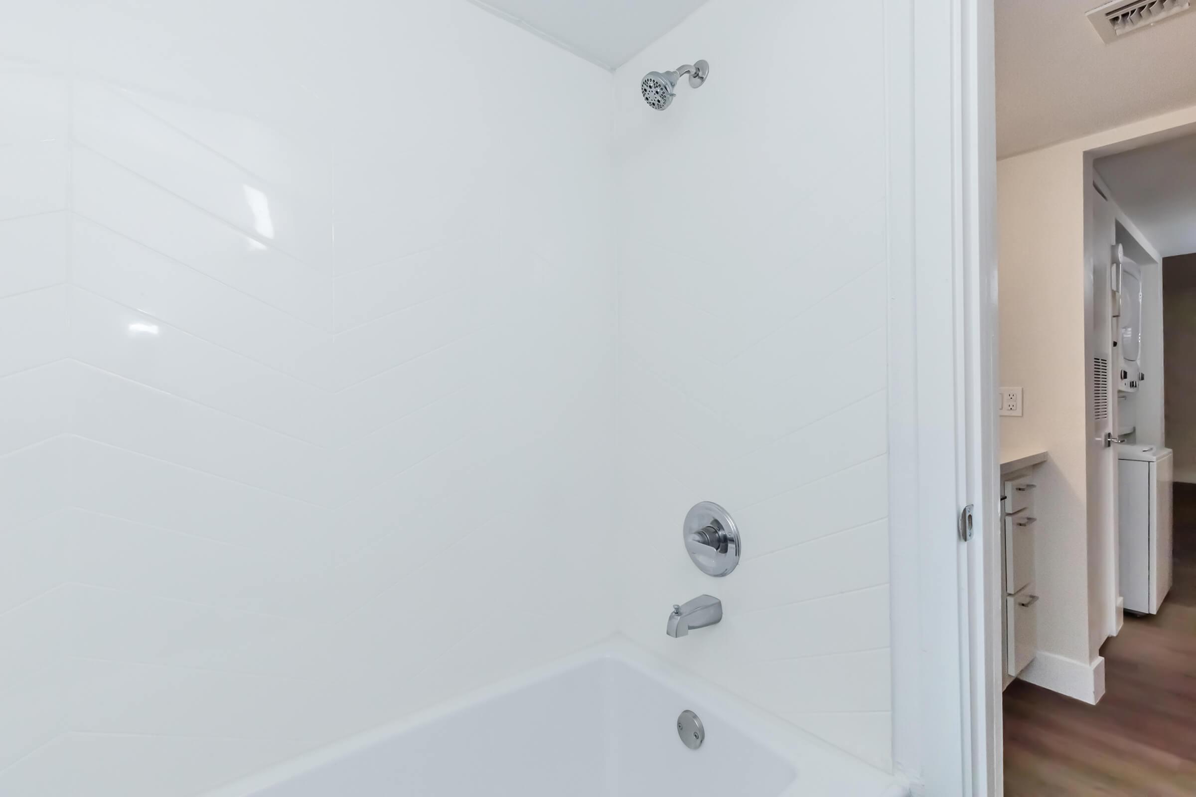 A clean, modern bathroom featuring a white tiled shower area with a showerhead and faucet. The bathtub is visible beneath the shower. The walls are tiled in a chevron pattern, and the background shows an unobtrusive glimpse of a hallway.