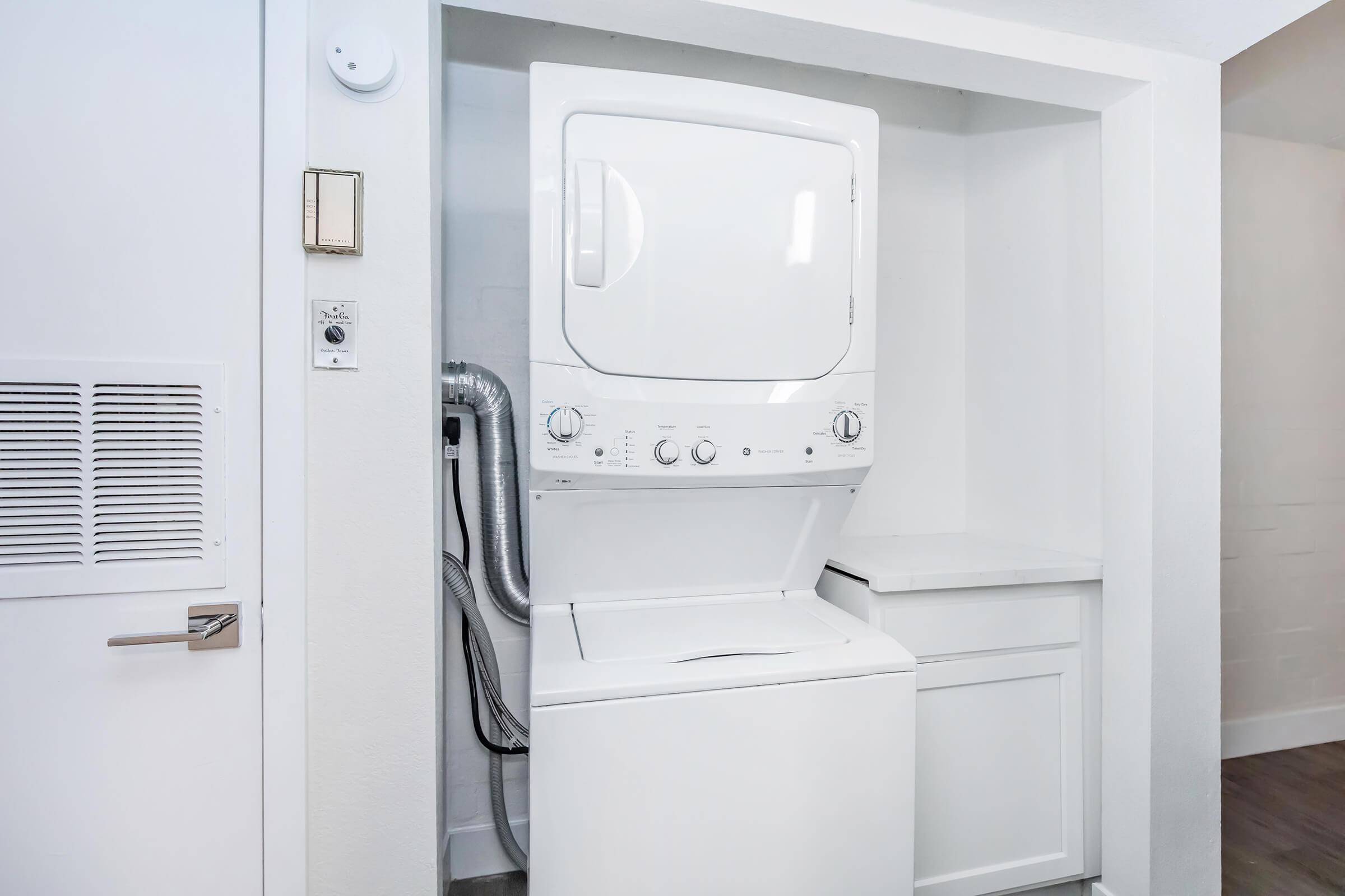 A stacked washer and dryer unit is installed in a compact laundry area. The machines are white and feature controls on the front. There is a small countertop next to the dryer, and the space is well-lit with a minimalist design against light-colored walls.