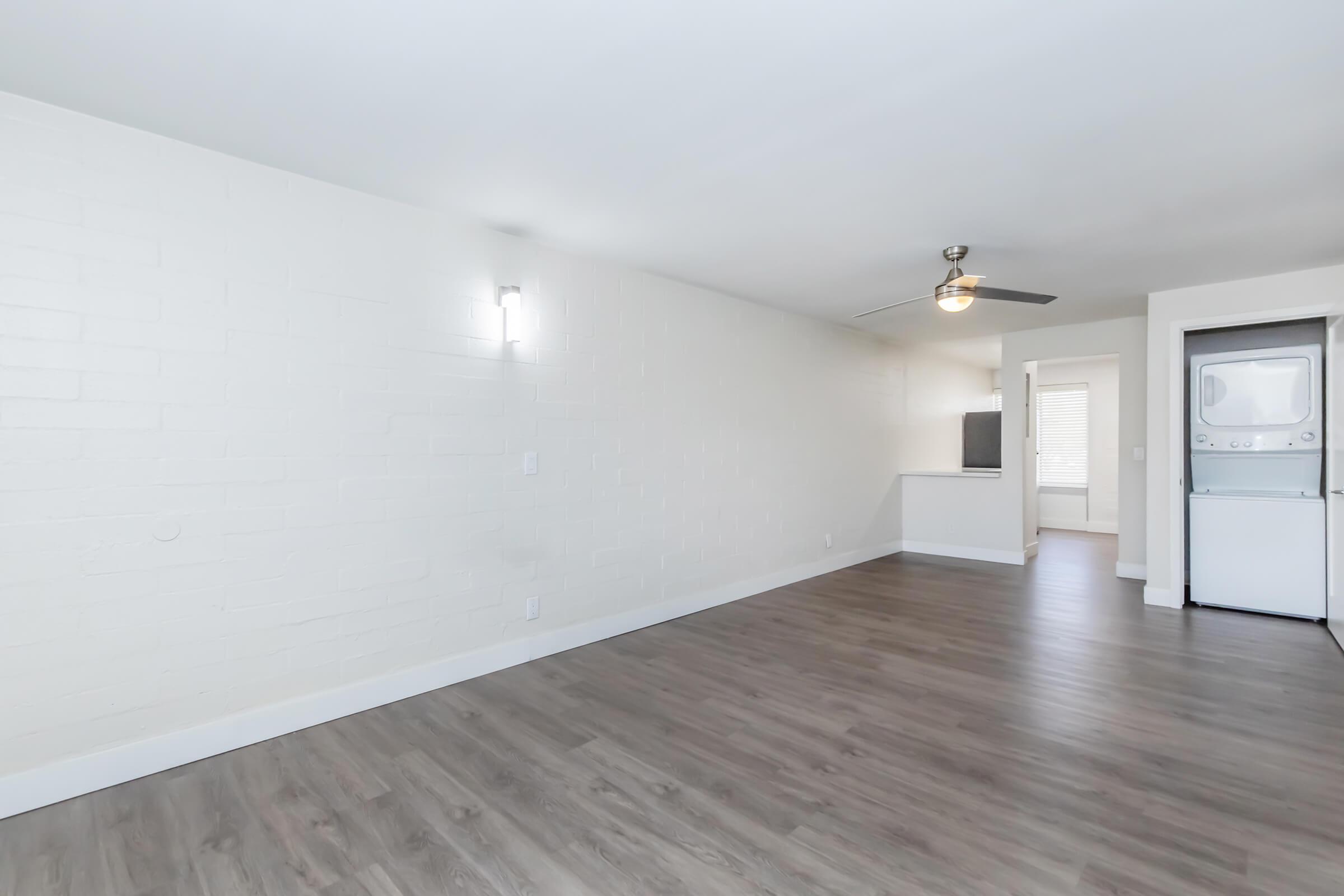 A spacious interior of an apartment featuring light-colored walls, wooden flooring, a ceiling fan, and modern lighting. In the background, there is a small kitchen area visible, along with a laundry space equipped with a washer and dryer. The room is bright and inviting, creating an open atmosphere.