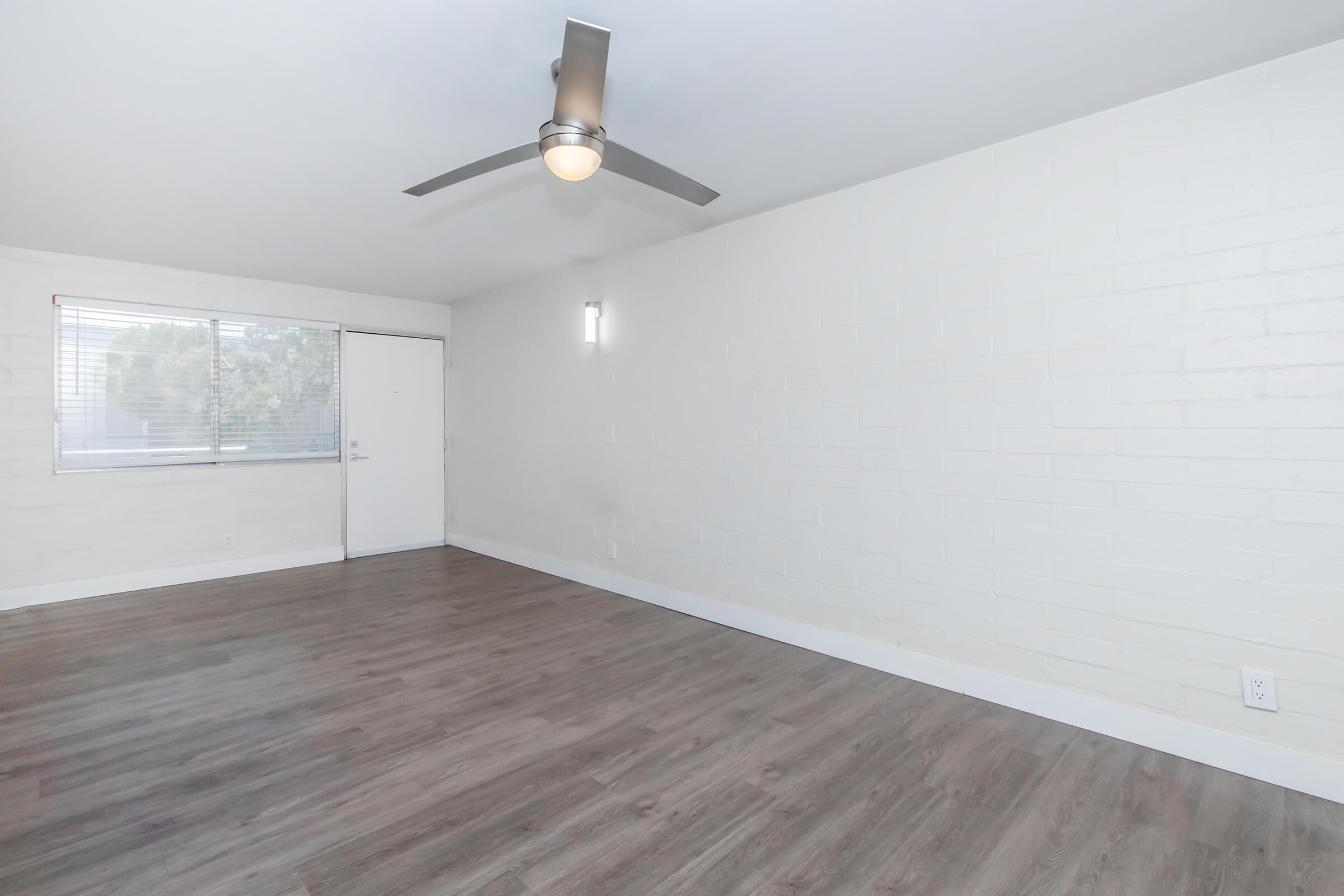 A spacious, minimally furnished room featuring light-colored walls, a ceiling fan, and a large window with natural light. The flooring is a dark wood finish, and there is a doorway leading to another area. The overall atmosphere is bright and inviting, suited for various design possibilities.