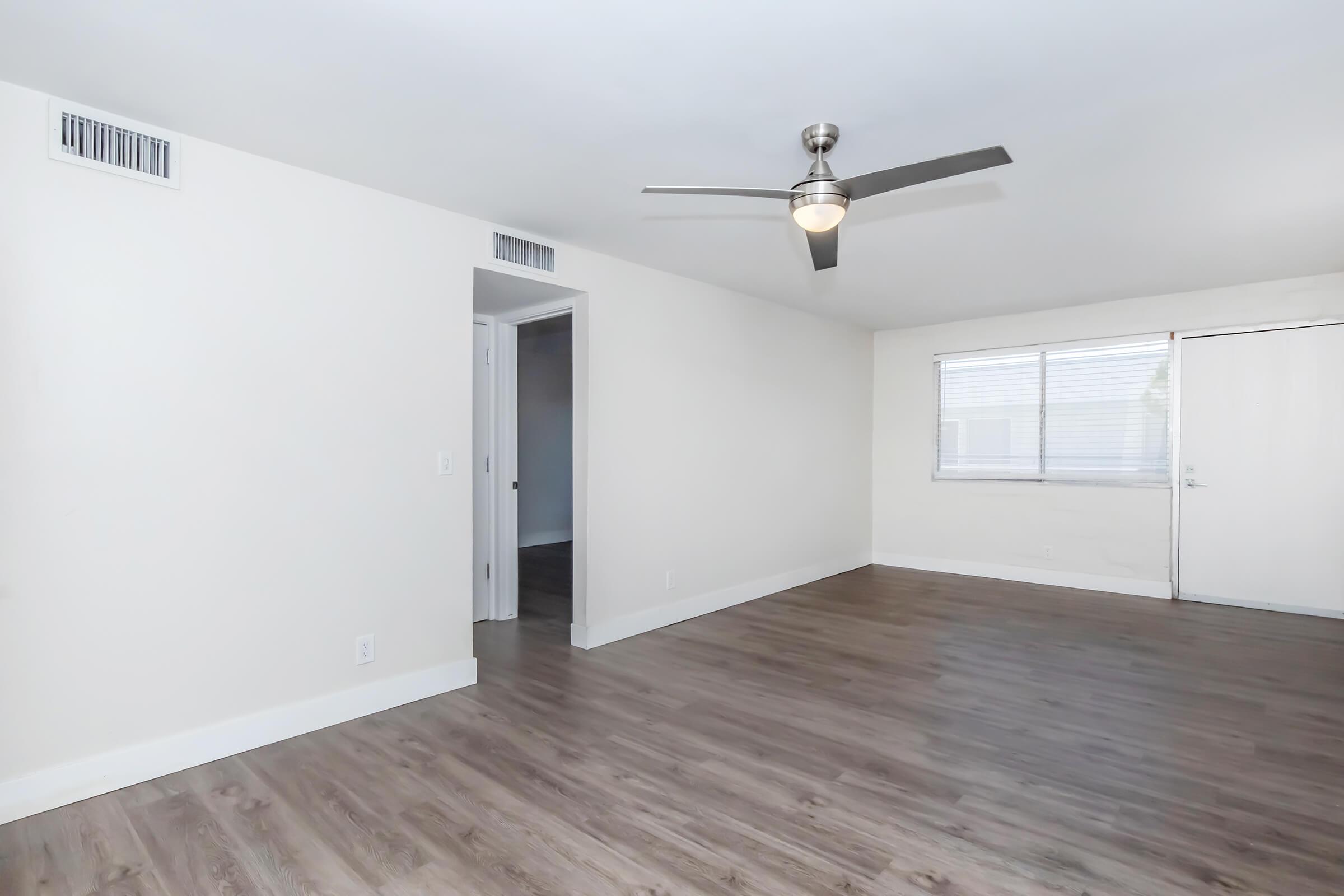 A spacious, well-lit room with light-colored walls, a ceiling fan, and wood-style flooring. There is a door leading to another room on the left and a large window on the right, allowing natural light to enter the space. The overall atmosphere is clean and modern.