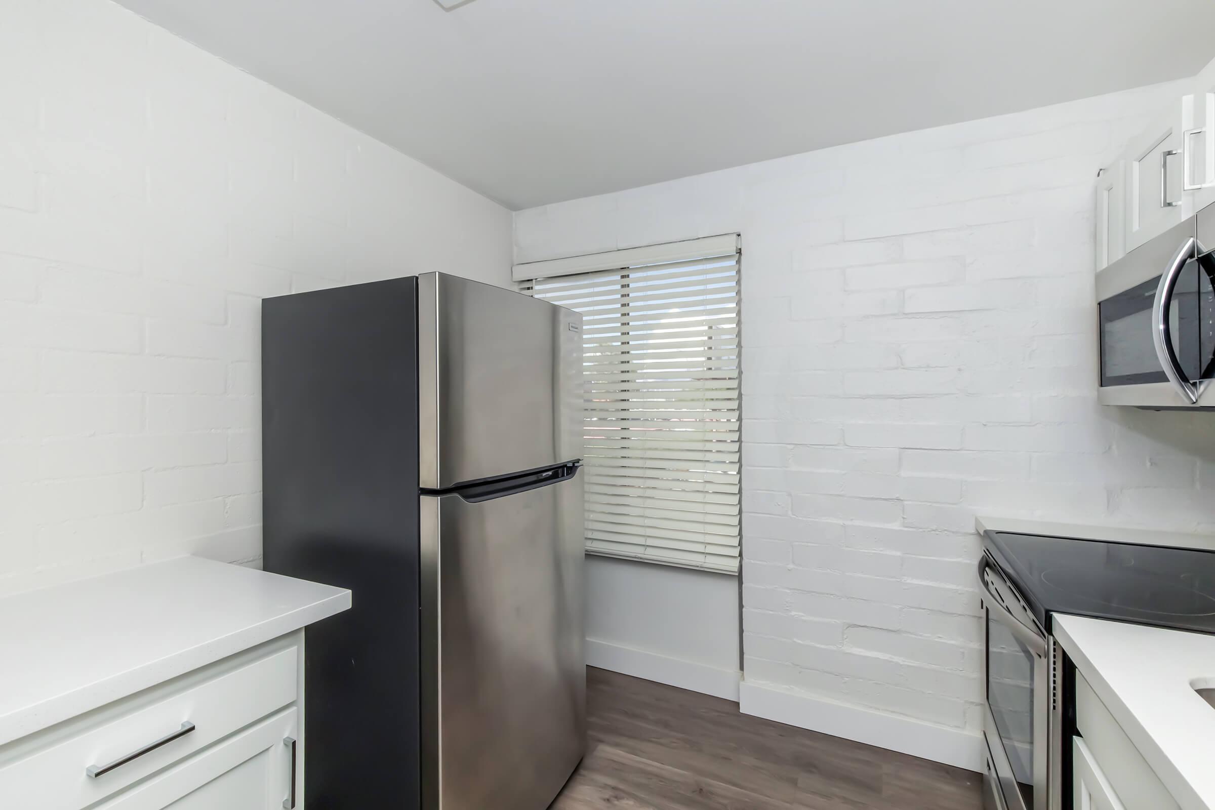 Modern kitchen featuring a stainless steel refrigerator, white brick walls, a window with blinds, and sleek cabinetry. The space is bright and minimalistic with a neutral color palette and contemporary appliances.