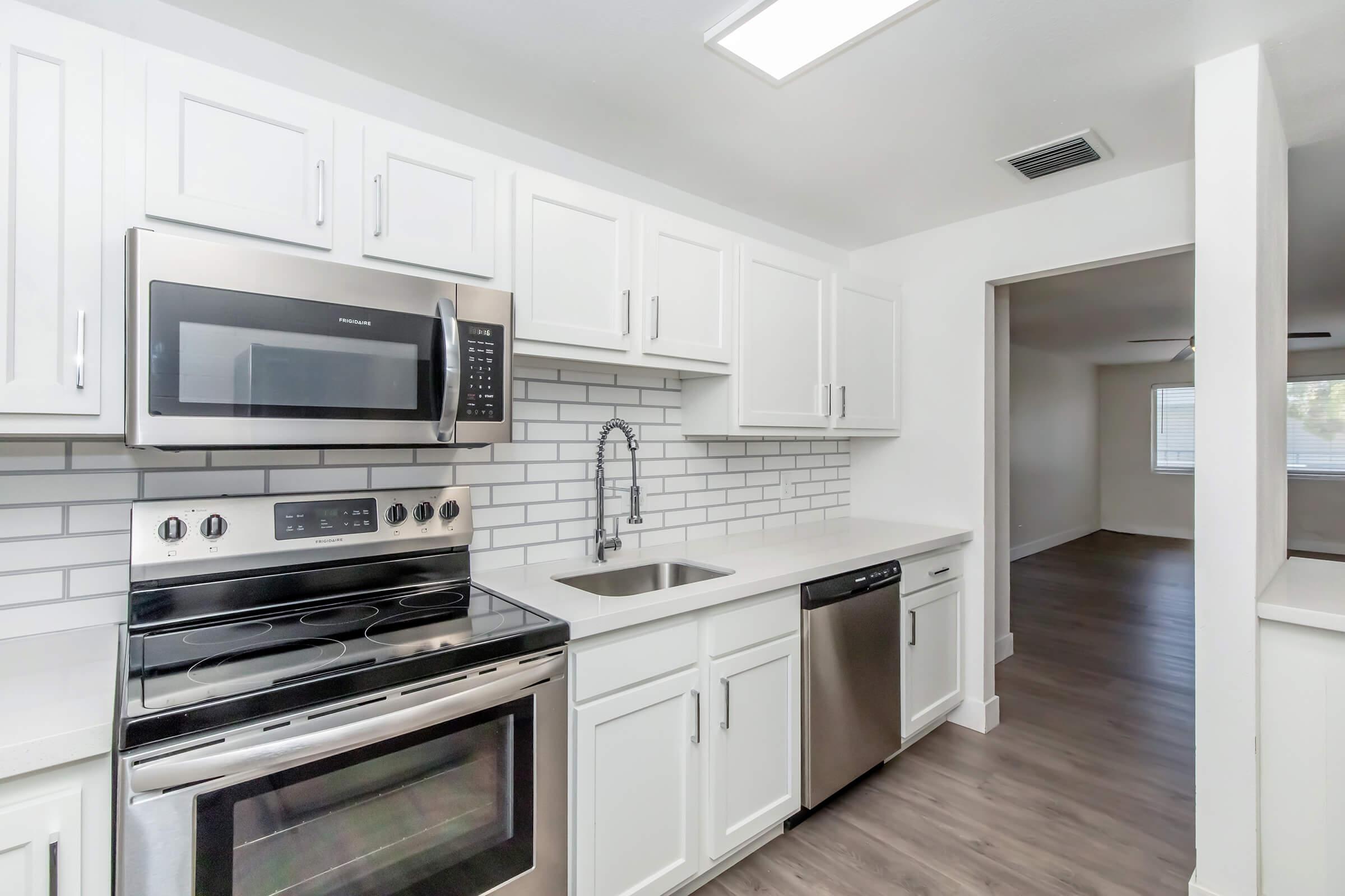 Modern kitchen featuring white cabinetry, stainless steel appliances including a microwave, oven, and dishwasher, and a sleek tiled backsplash. The countertop is light-colored. A doorway in the background leads to a spacious living area with natural light.