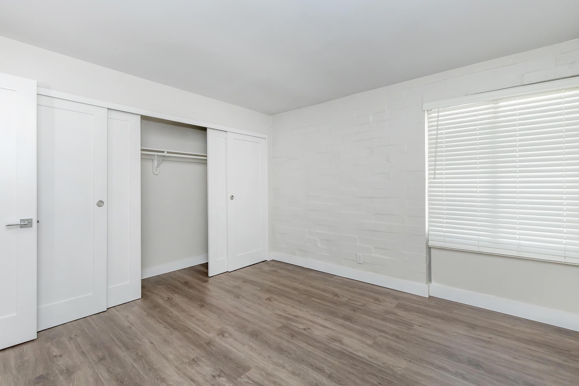 An empty room with light-colored walls, featuring a closet with sliding white doors on the left and a window with white blinds on the right. The floor has a wood-like finish, adding warmth to the space.
