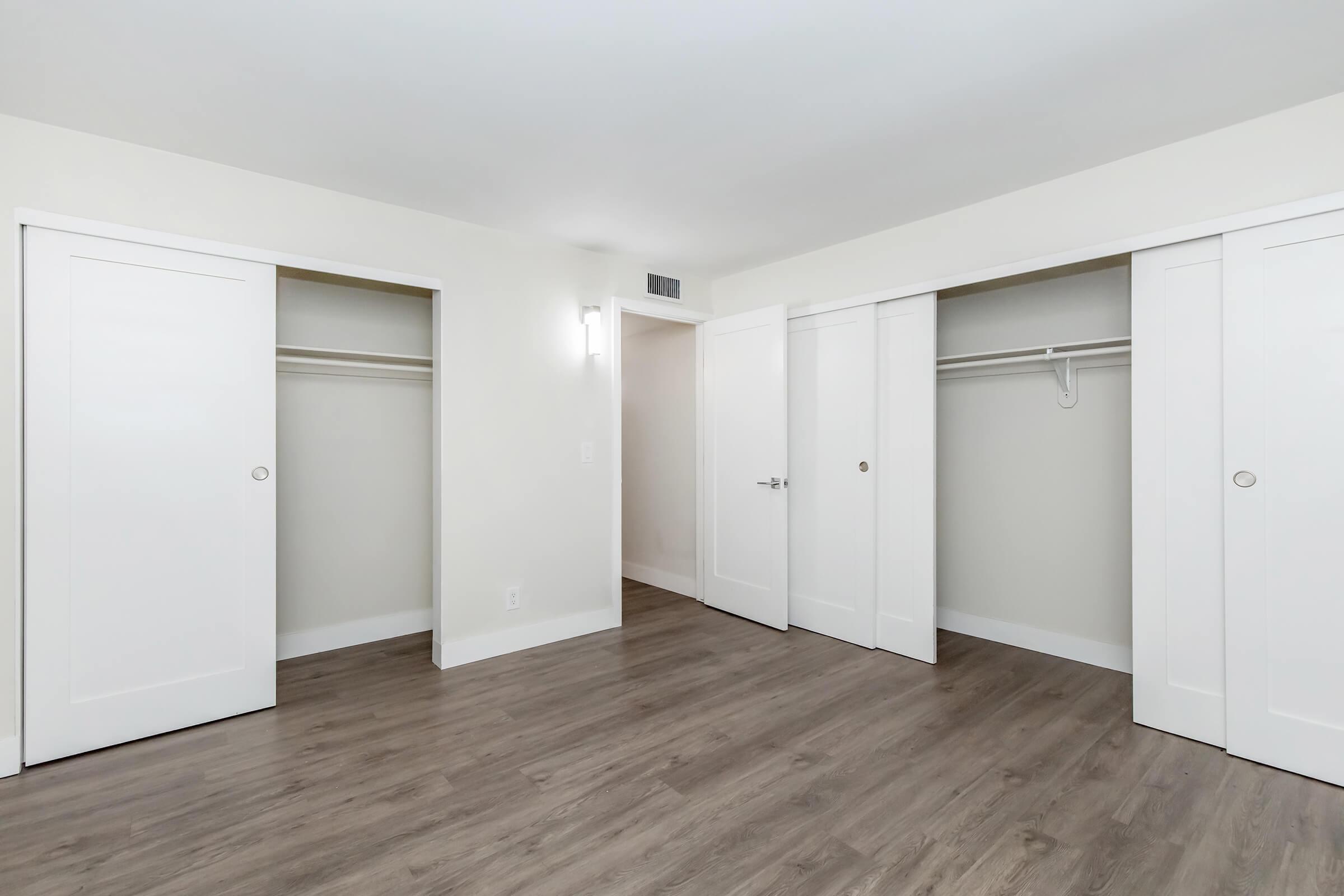 A well-lit, empty room featuring two sliding door closets with white doors. The walls are painted a light color, and the floor is a dark wood laminate. The room has a modern feel, with clean lines and minimalistic design, providing a spacious and uncluttered appearance. 