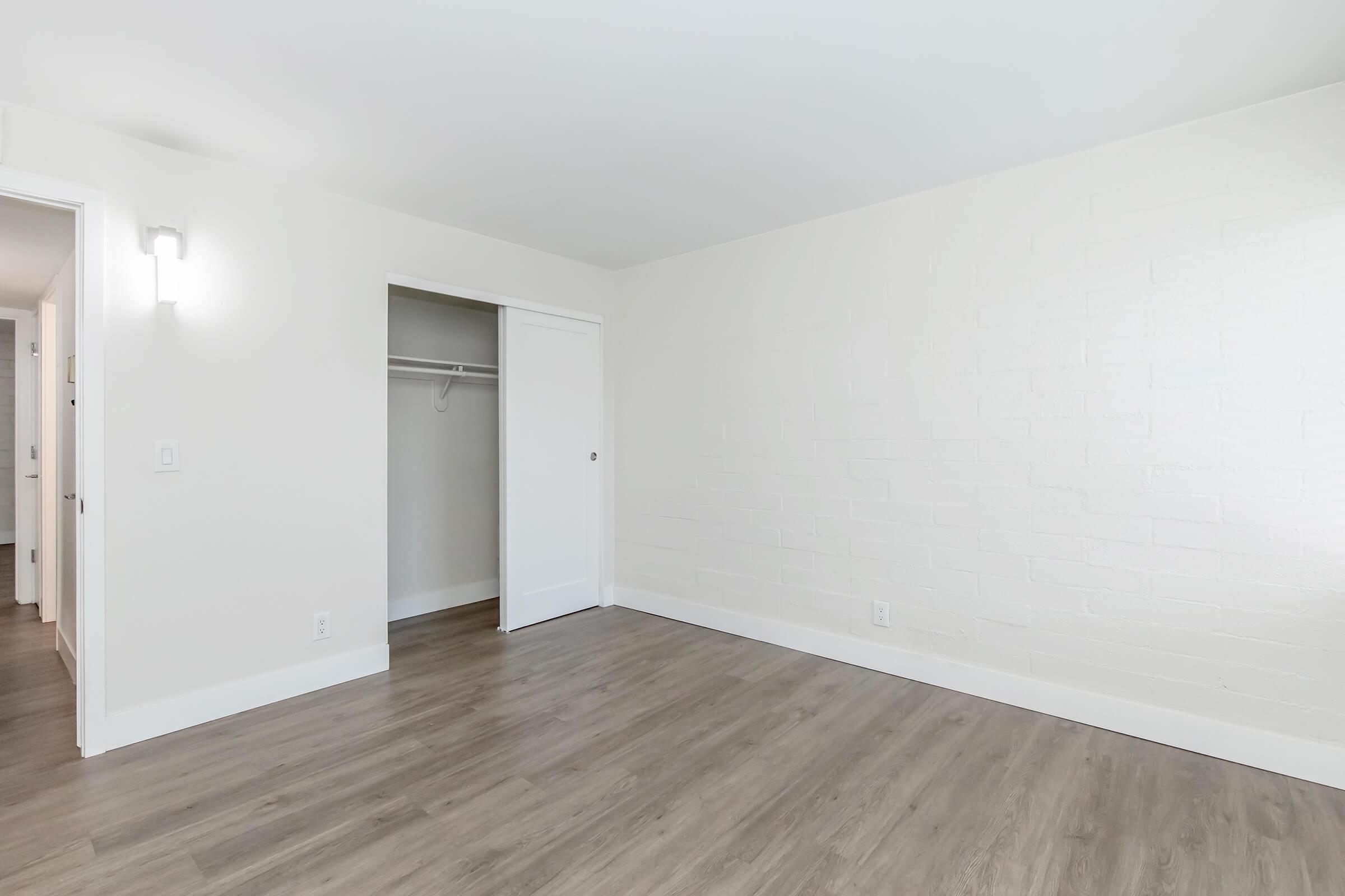 A spacious, empty room with white walls and light wood flooring. There is a closet with sliding doors on the left and a wall-mounted light fixture. The room has a bright and airy feel, with no furniture or decorations present.