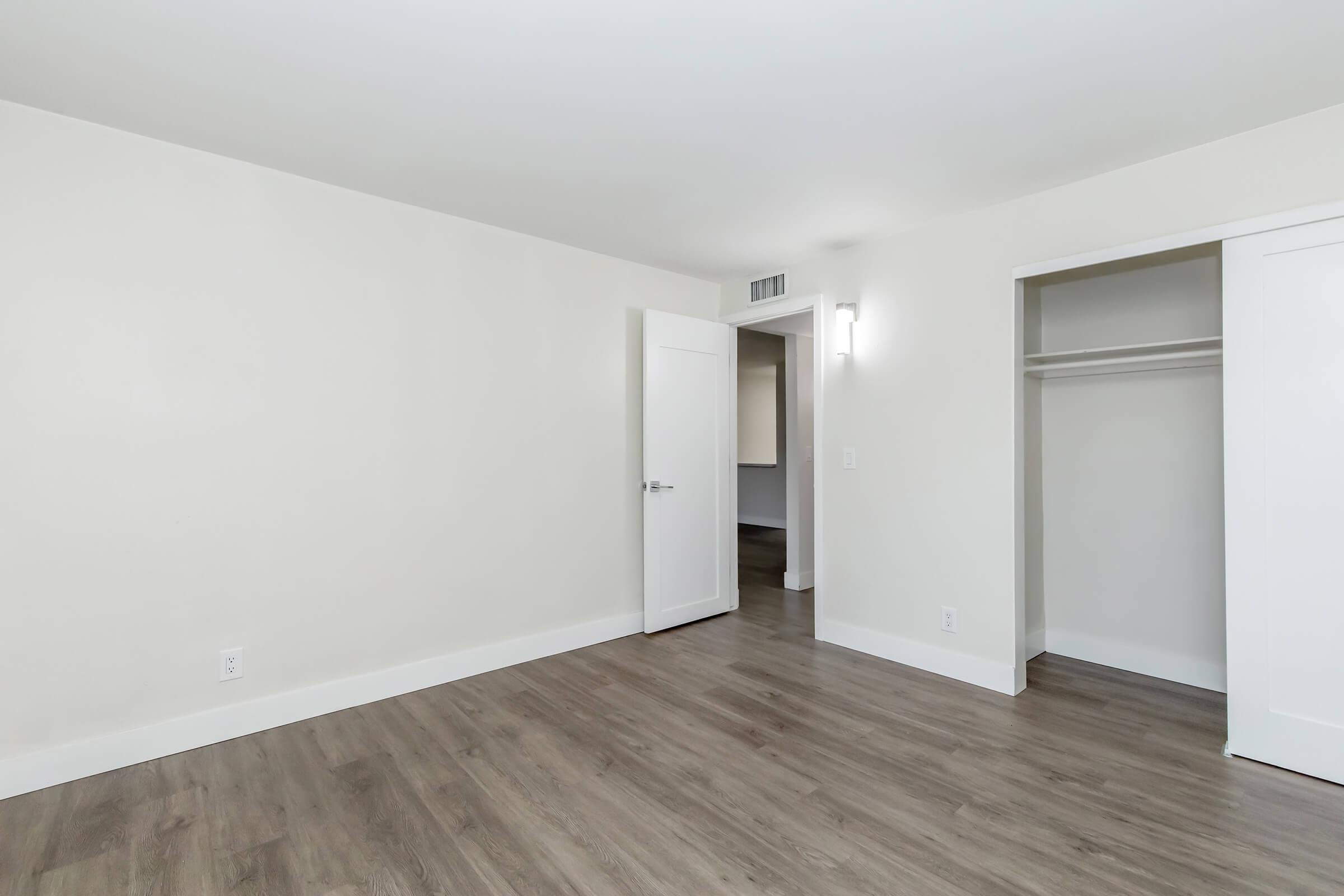 A bright, empty room with light-colored walls and hardwood flooring. There is a white closet with sliding doors on one side, and a door leading to another room. The space is well-lit with a modern wall sconce. The overall atmosphere is clean and minimalistic.