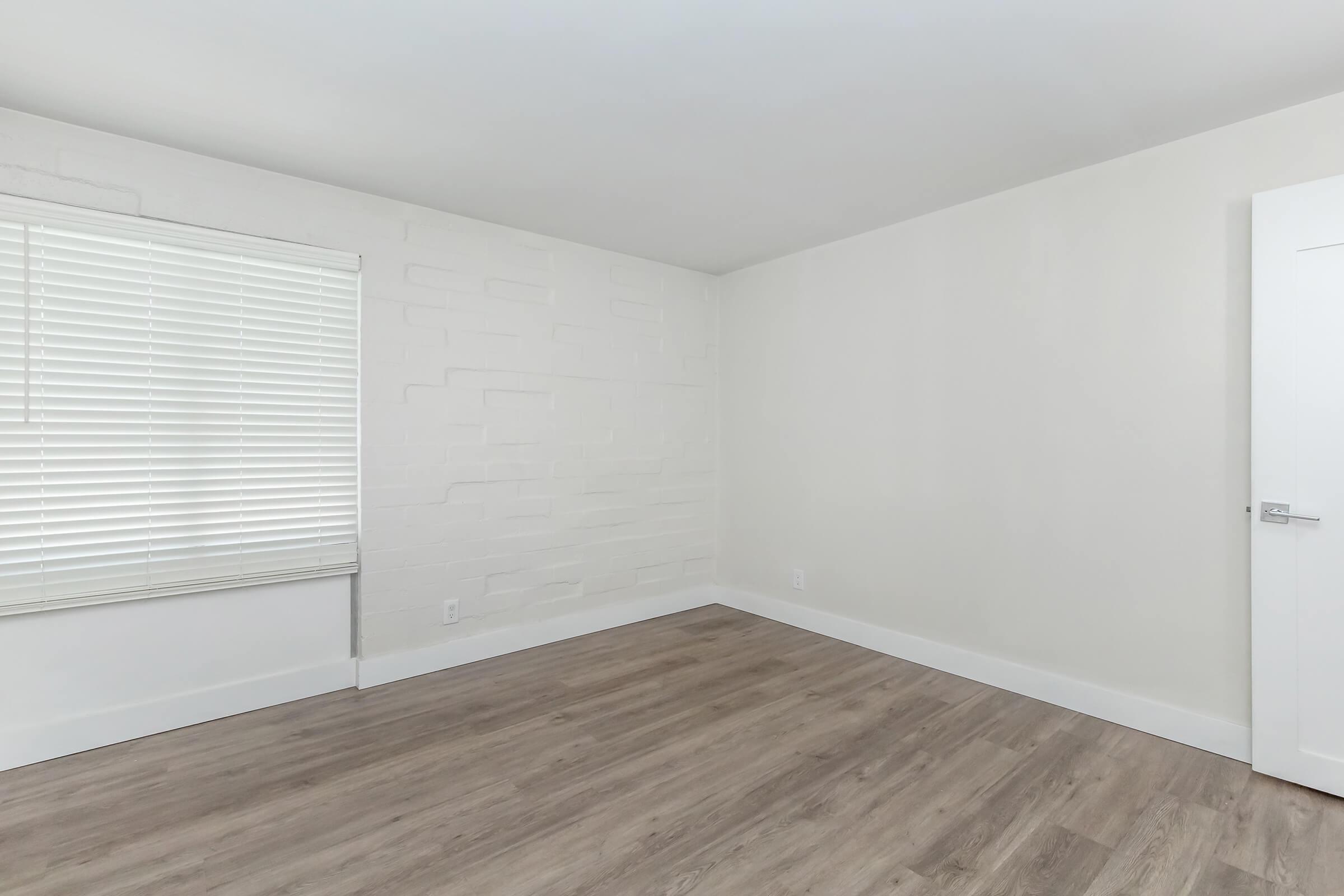 A spacious, modern room featuring a light-colored, textured wall, large window with white blinds, and laminate wood flooring. The room has a clean, minimalistic design with ample natural light and a door on the right leading to another space.