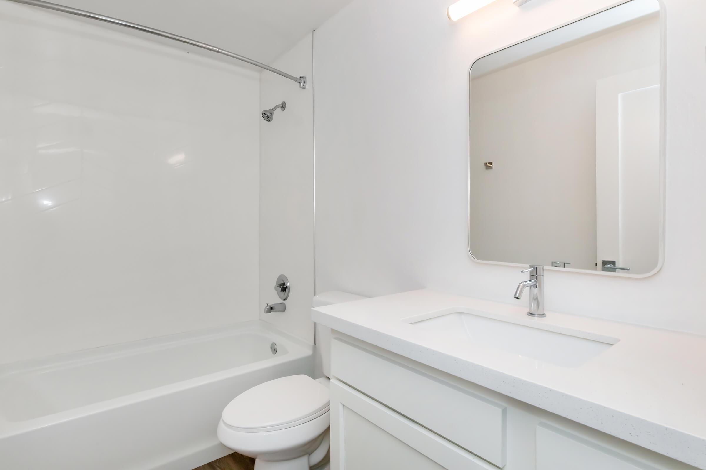 A modern bathroom featuring a white bathtub, shower with glass panel, a sleek white vanity with sink, and a rectangular mirror above it. The walls are painted white, and the flooring appears to be a light wood laminate. Bright and clean atmosphere with minimalistic design.