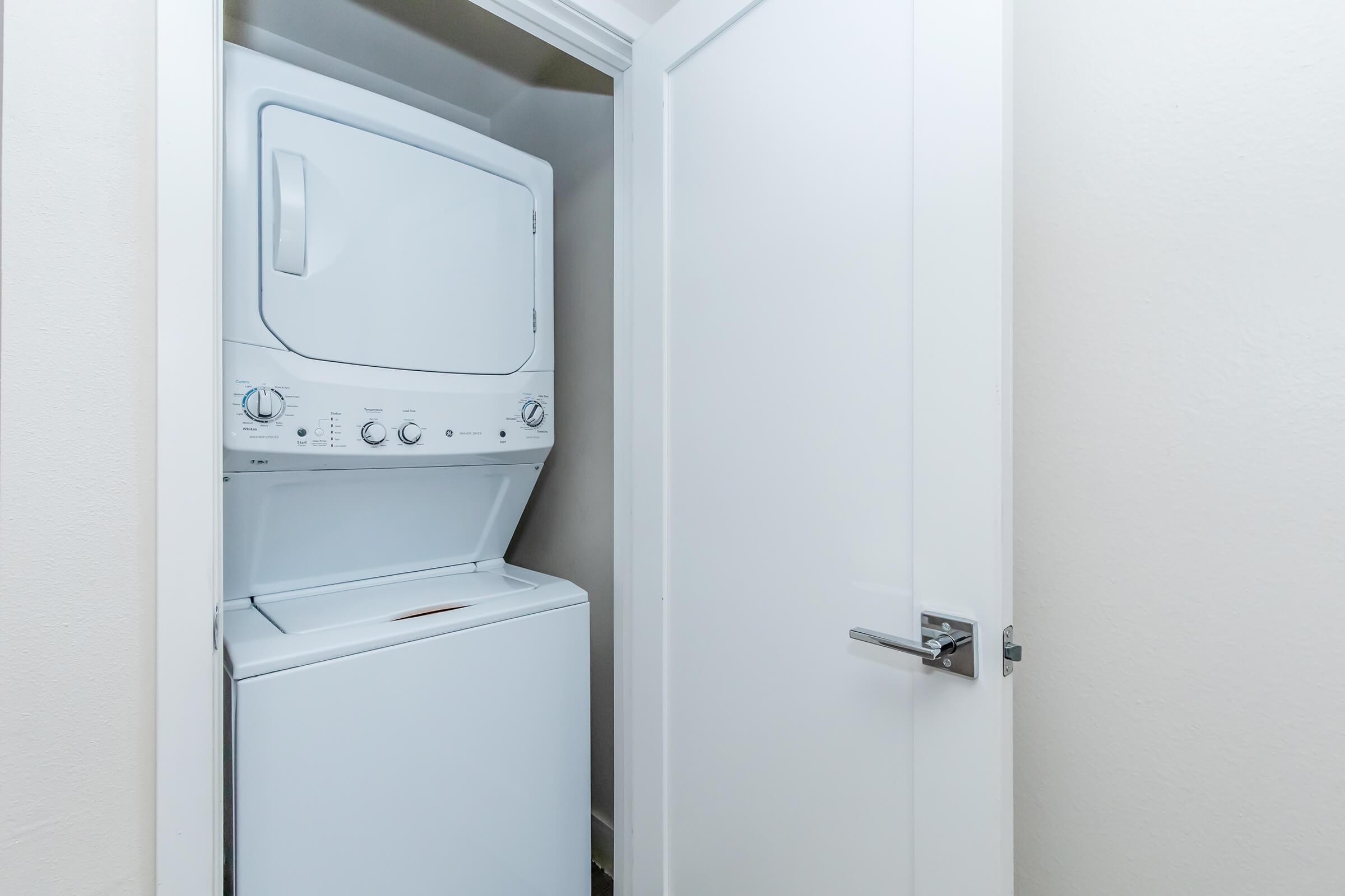 A compact laundry area featuring a stacked washer and dryer set. The appliances are white and placed inside a small closet space. A closed door is visible next to the appliances, and the walls have a light, neutral color.