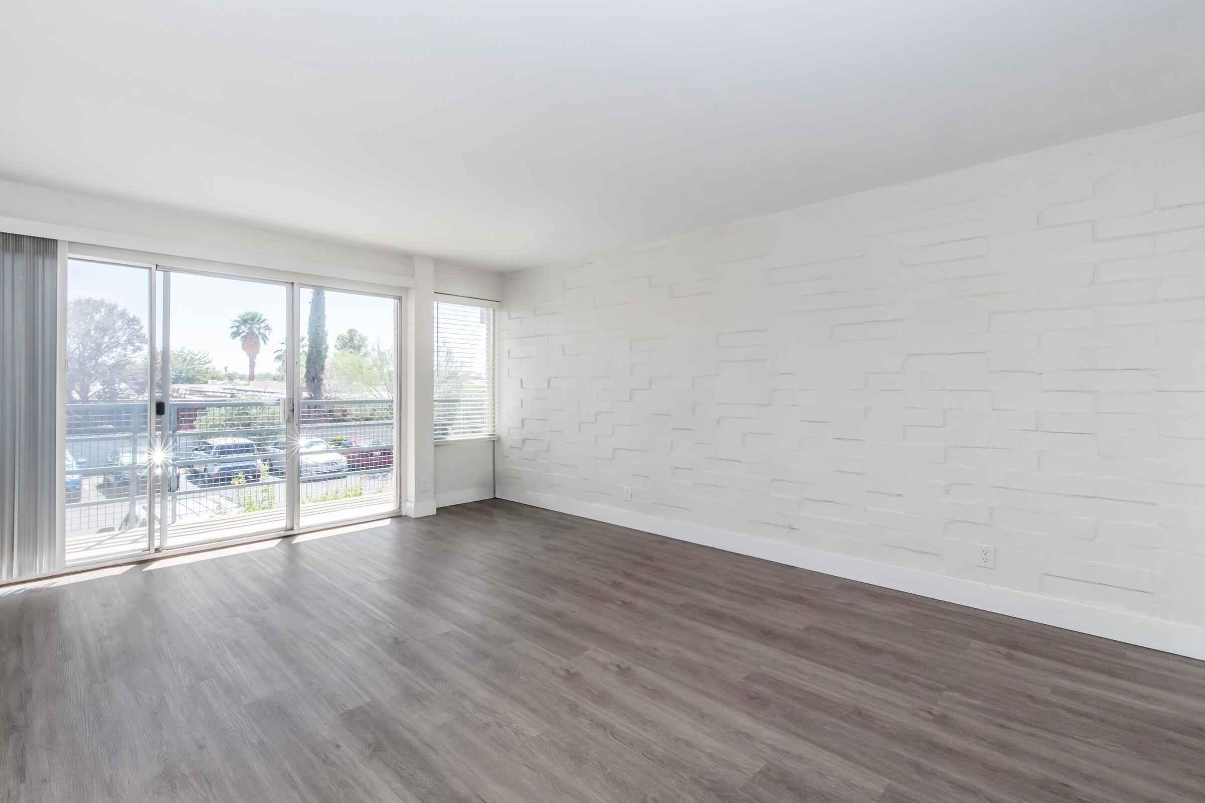 Empty room with a light-colored wall featuring a textured design, large windows allowing natural light, and a view of greenery outside. The floor is covered with light brown laminate flooring, creating a bright and spacious atmosphere. No furniture is present, emphasizing the room's openness.