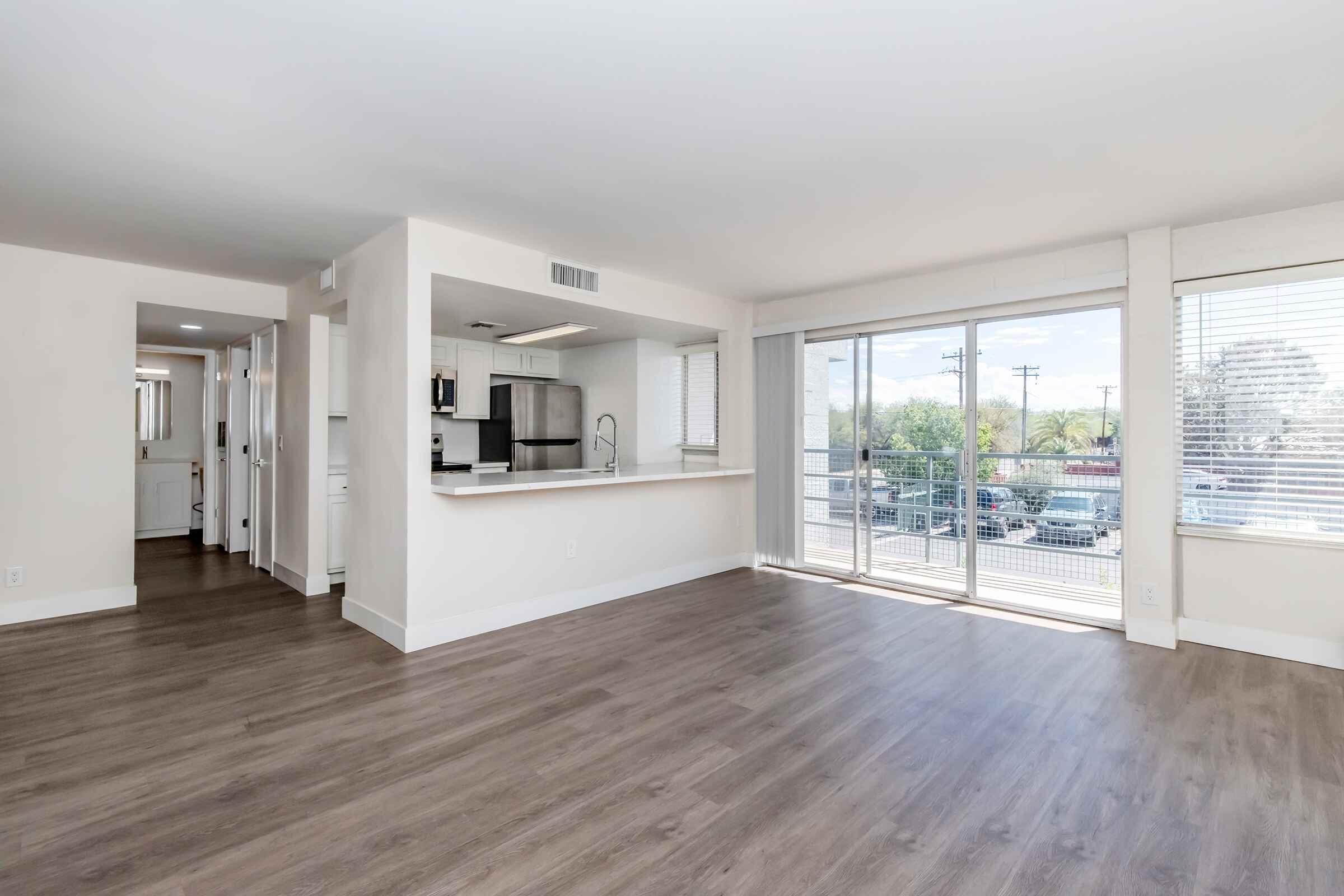 Spacious, modern apartment interior featuring a living area with large windows allowing natural light, a kitchen with an island bar, and a balcony view. The flooring is light wood, and the walls are painted in neutral tones, creating a bright and inviting atmosphere.