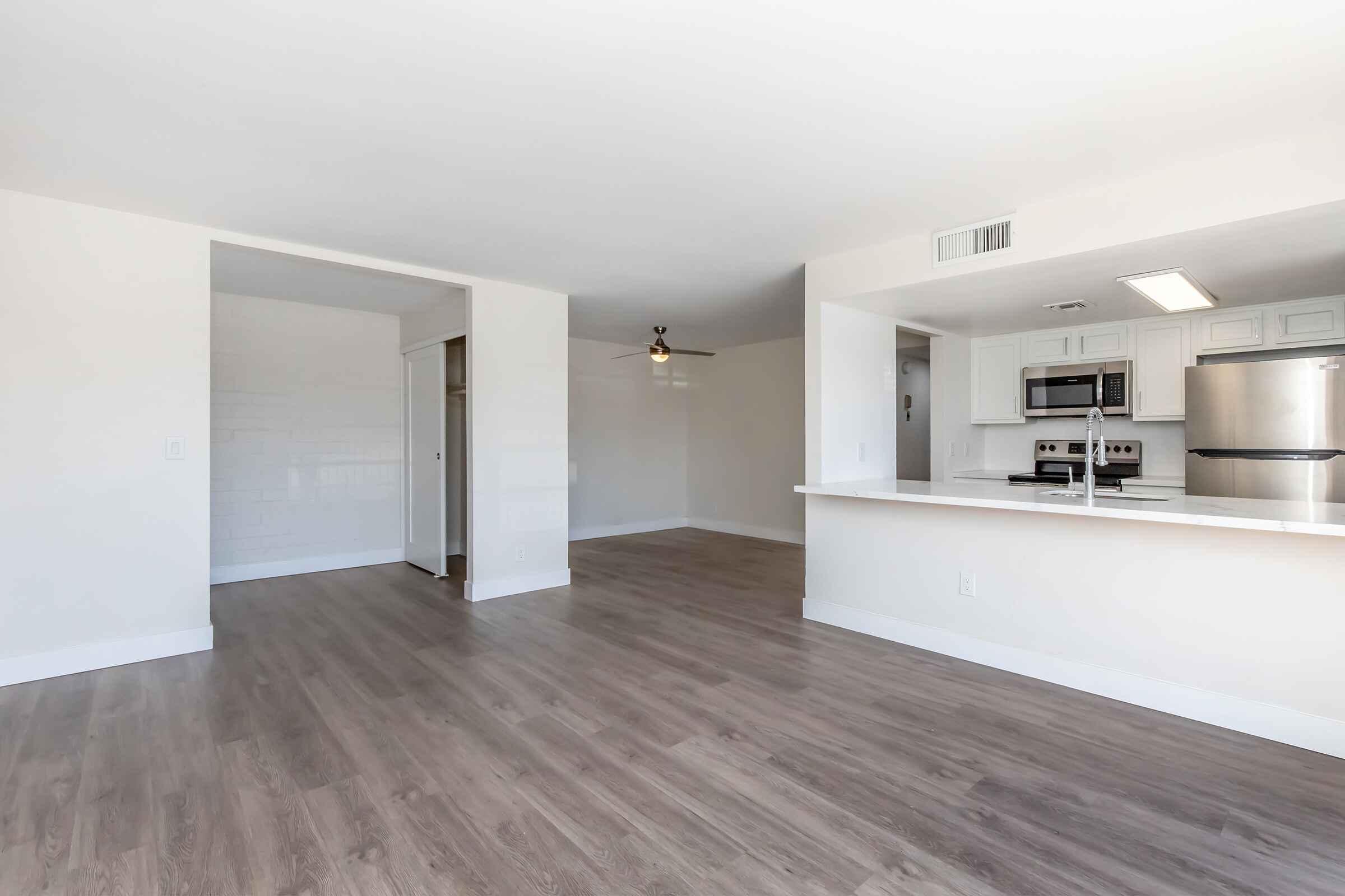 Interior view of a modern living space with an open floor plan. The area features light-colored walls, large windows allowing natural light, and light hardwood flooring. A kitchen area with stainless steel appliances is visible, along with a ceiling fan in the main room and a doorway leading to another space.
