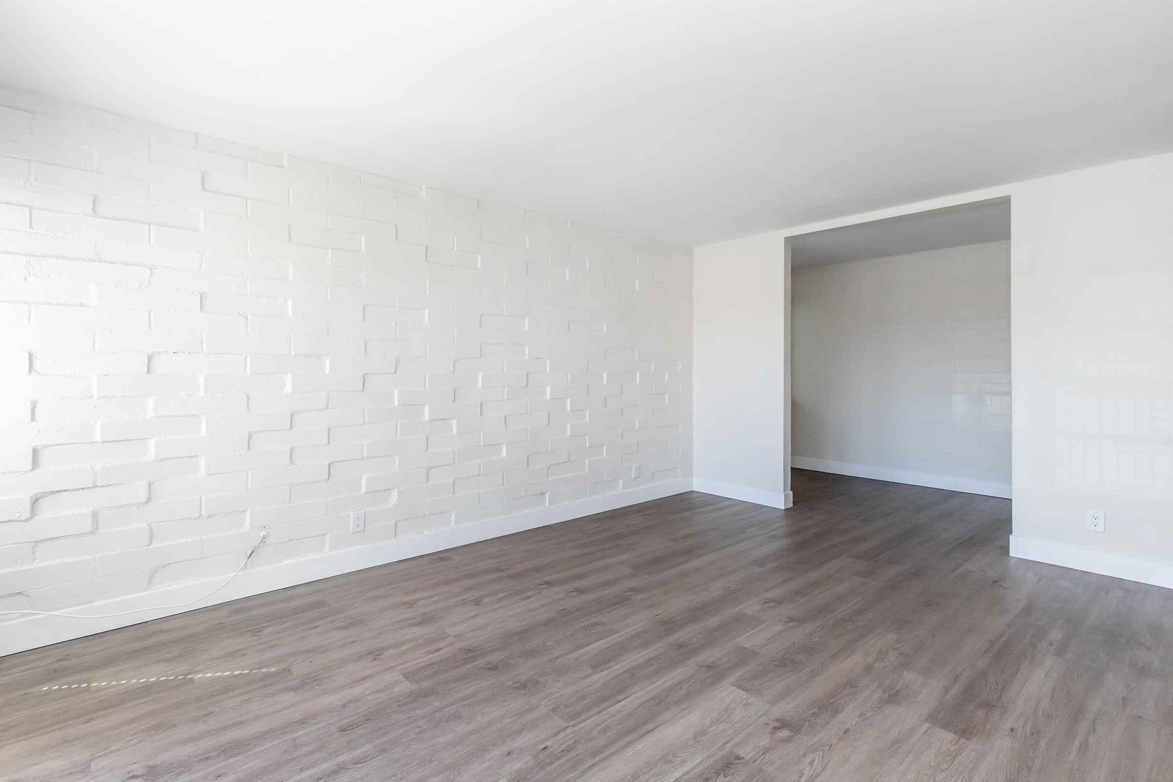 A spacious, empty room featuring a white brick wall and light-colored wooden flooring. The room has minimalist decor, with ample natural light and an opening leading to another area, creating a bright and inviting atmosphere.