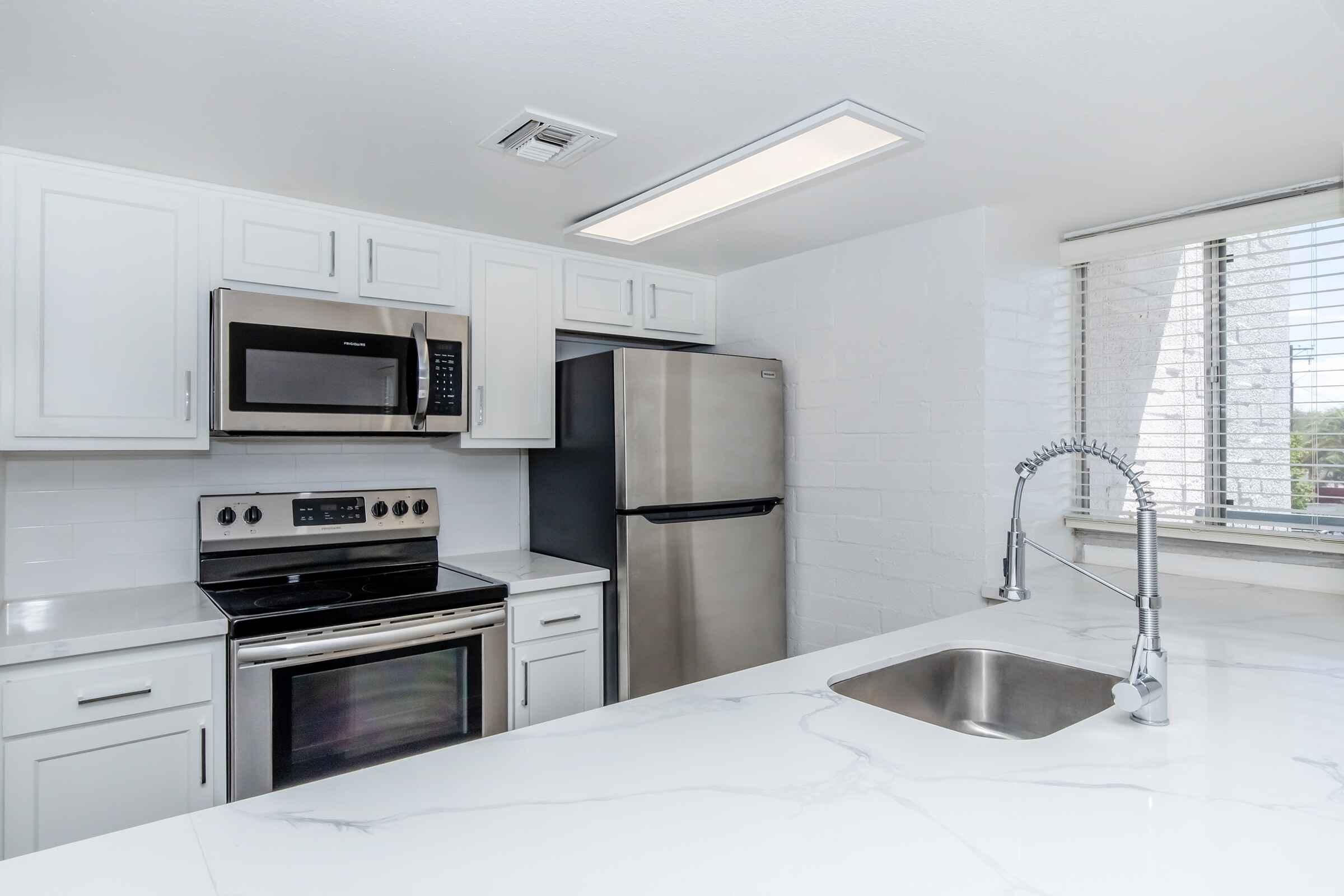 A modern kitchen featuring white cabinetry, stainless steel appliances including a microwave, oven, and refrigerator, and a marble countertop with a sink. Natural light enters through a window adorned with blinds, highlighting the clean and sleek design of the space.