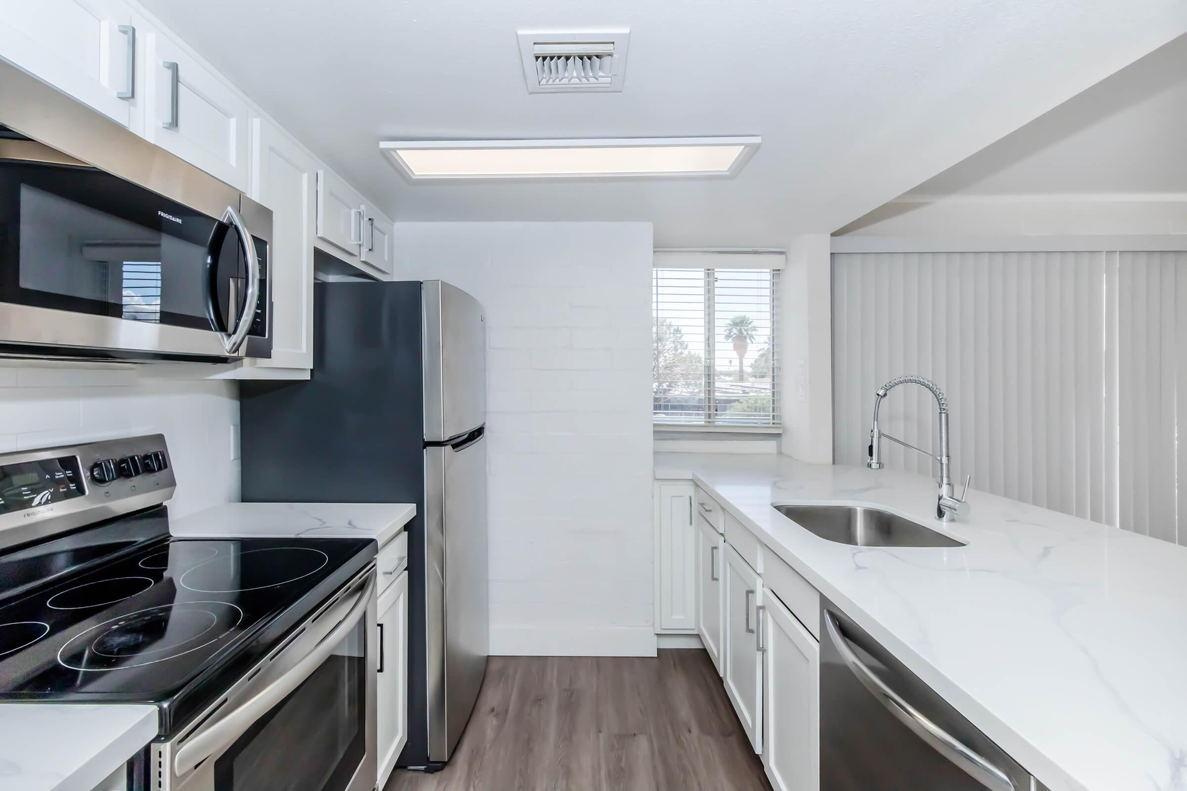 Modern kitchen with stainless steel appliances including an oven, microwave, and refrigerator. Features white cabinetry, a marble countertop, and a sleek sink. The room is bright with natural light coming through a window, and there are blinds installed for privacy.