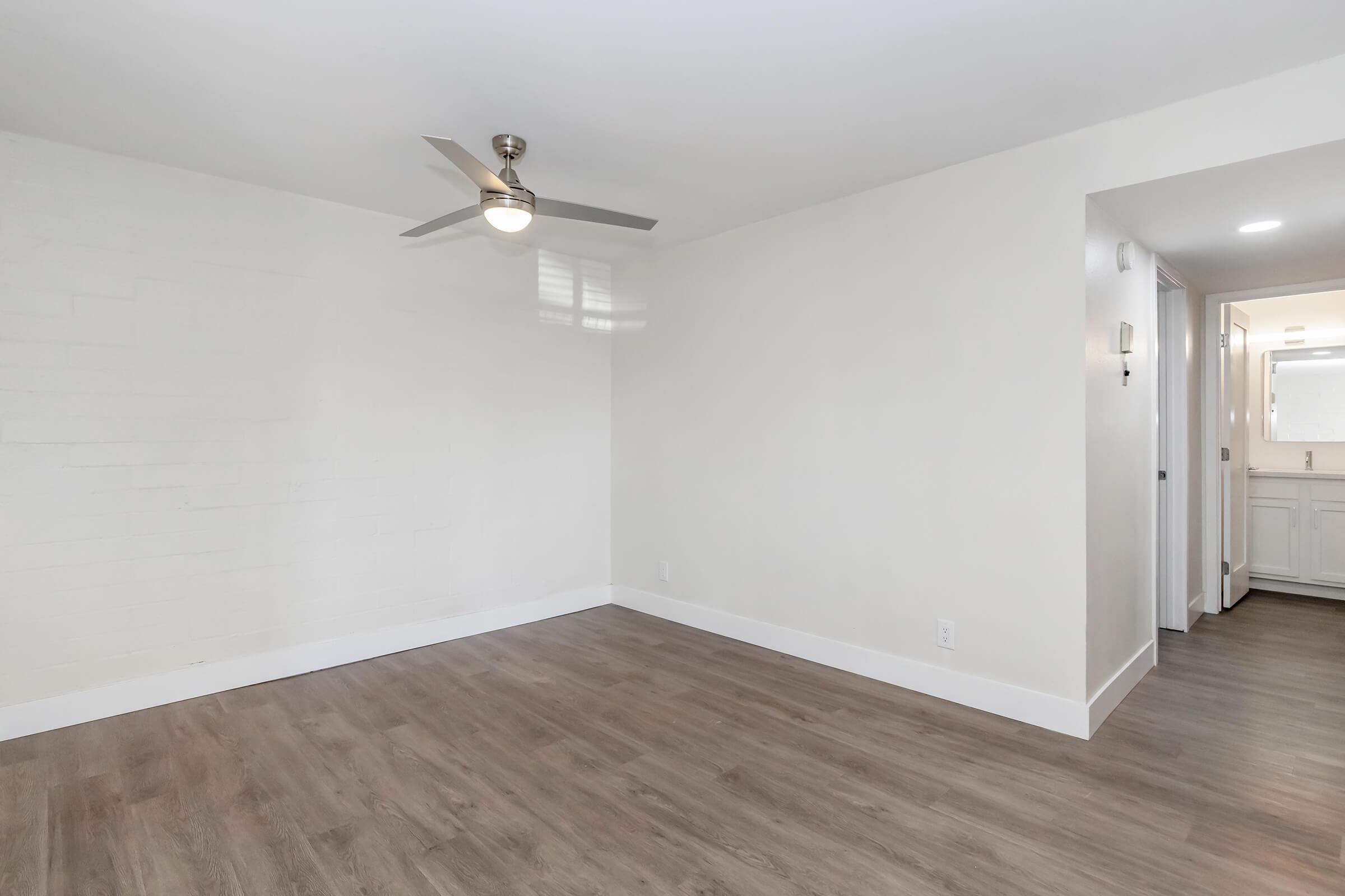 A spacious, well-lit room featuring light-colored walls and wooden flooring. A ceiling fan is installed, and there is a doorway on the right leading to a bathroom area, which is softly illuminated. The space has a clean, modern aesthetic.