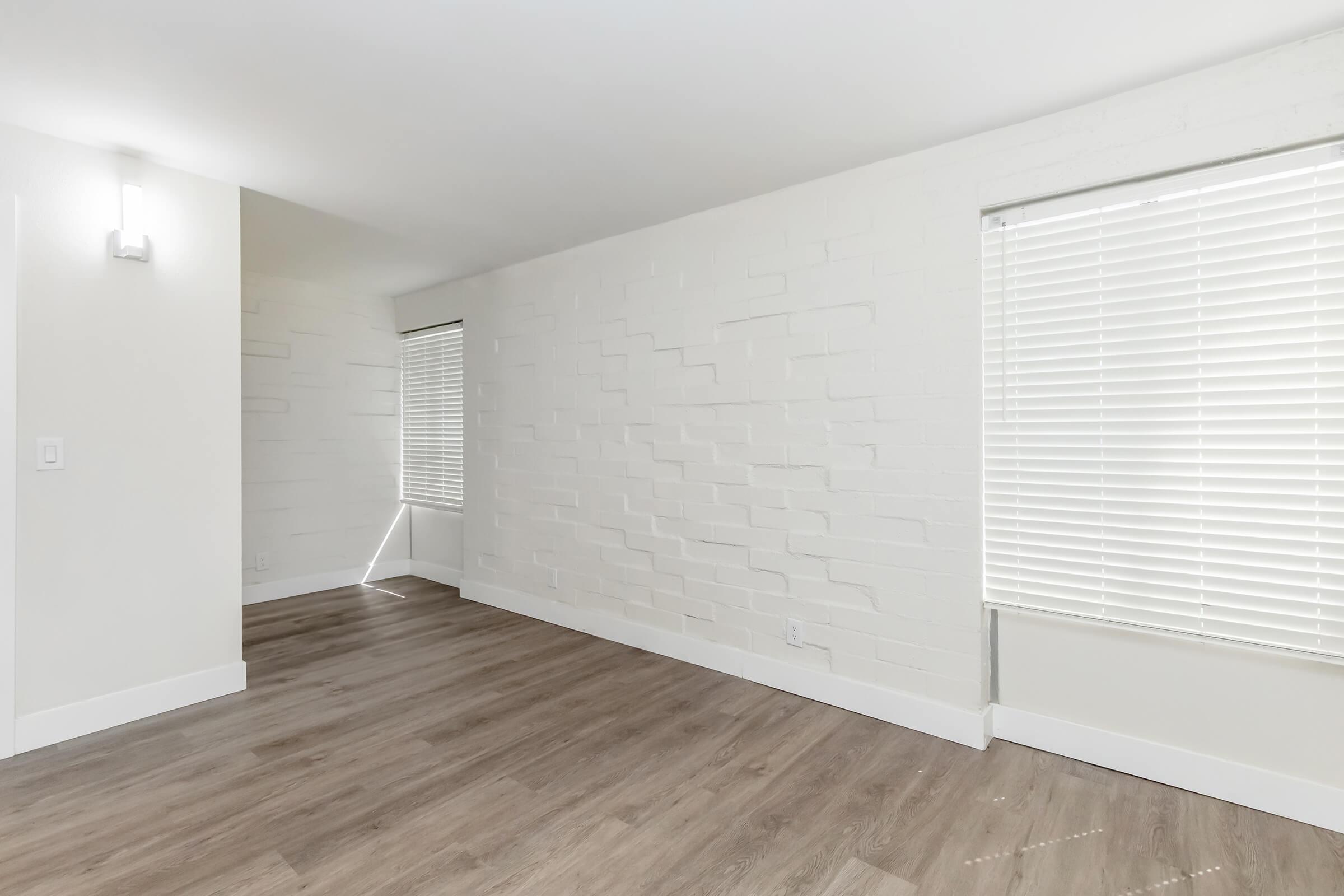 A bright, minimalist room featuring a textured white brick wall, large windows with blinds, and light wooden flooring. The space has a clean and modern aesthetic with ample natural light.