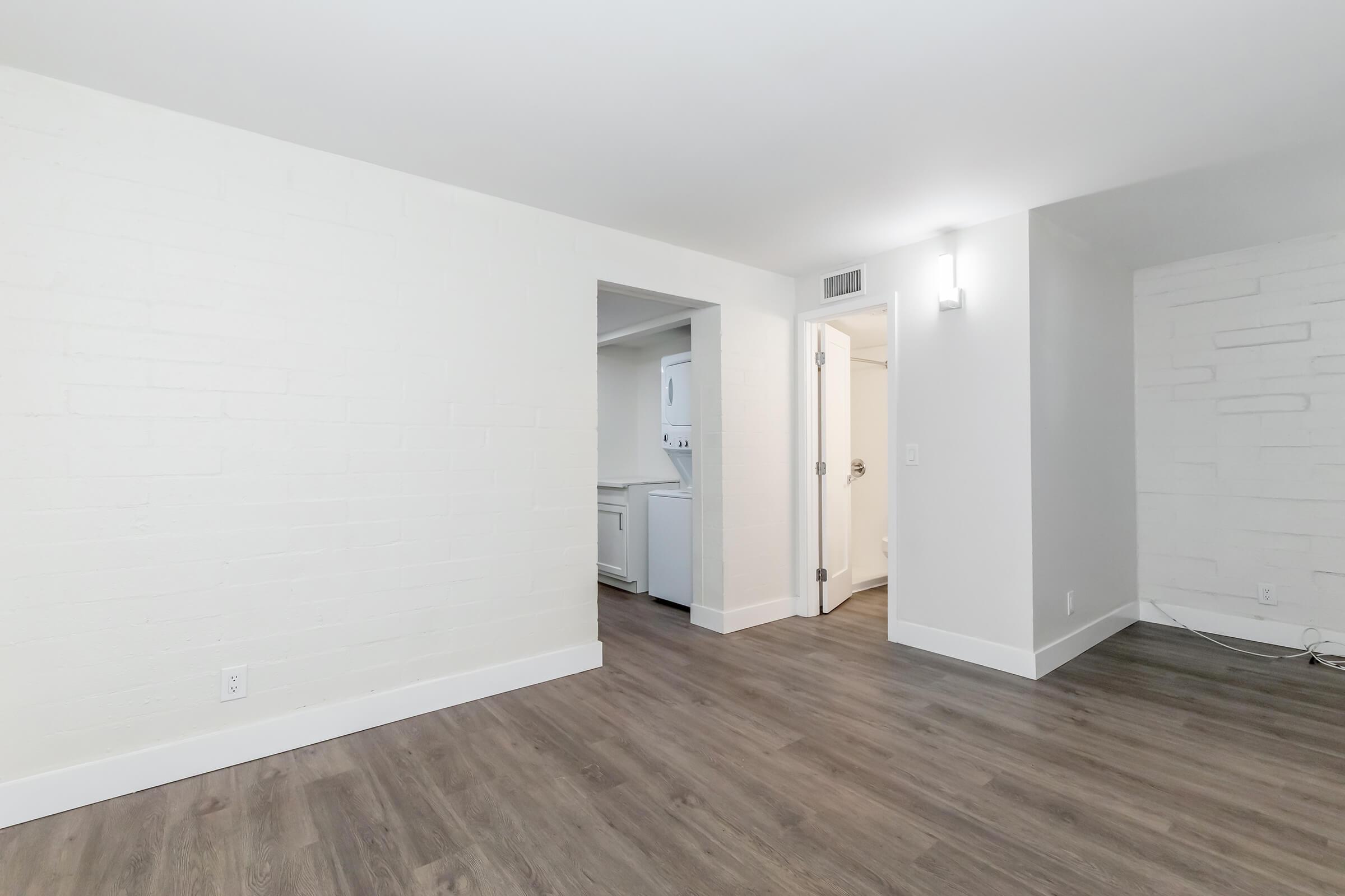 A bright, modern interior of a room featuring white walls, a wood-style floor, and a doorway leading to a kitchen area. The room has minimalist decor and ample natural light. A second doorway is visible, leading to a bathroom. The overall aesthetic is clean and contemporary.