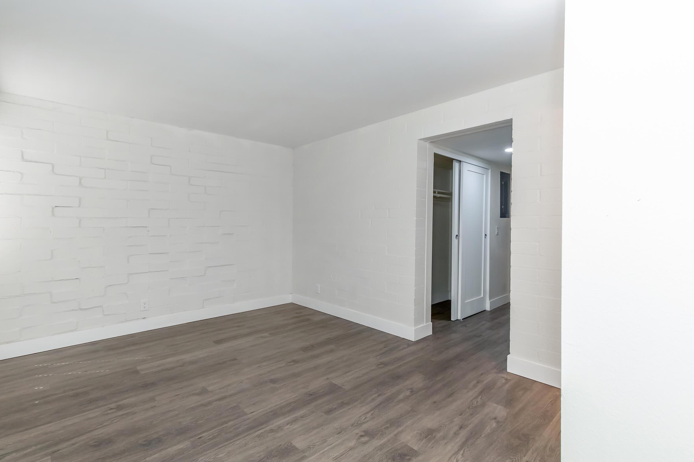 An empty room with white walls, featuring a textured wall on the left side. The floor is covered with light brown laminate. A doorway on the right leads to a hallway with another room visible. The overall atmosphere is bright and minimalistic.