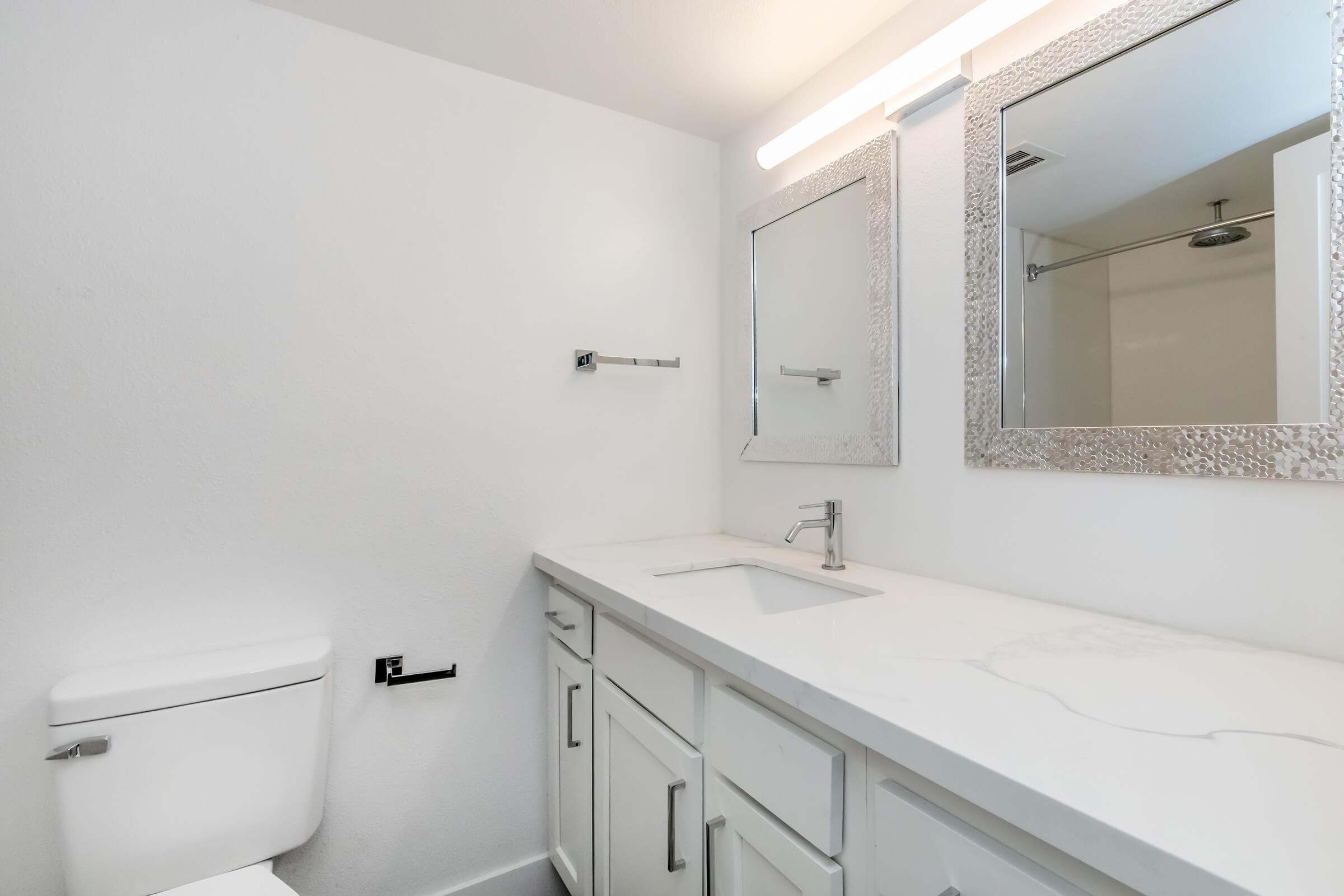 A modern bathroom featuring white walls, a clean white toilet, and a sleek vanity with a sink. There are two rectangular mirrors above the vanity, framed in a textured silver design. A light fixture is mounted above the mirrors, illuminating the space. The overall aesthetic is bright and minimalistic.