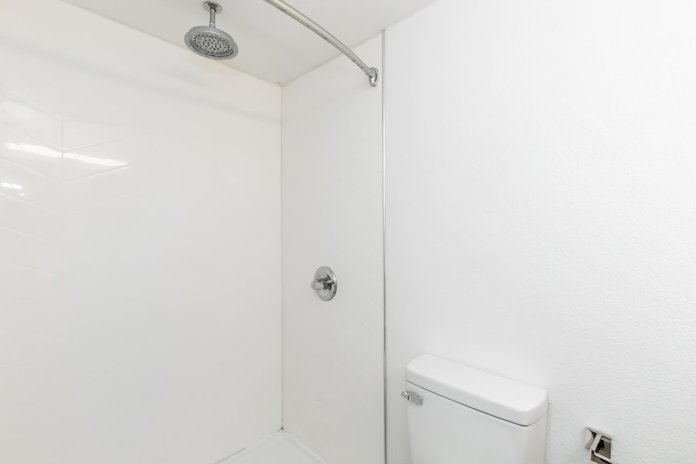 A clean bathroom featuring a shower area with a modern showerhead and a toilet. The walls are smooth and white, creating a bright and minimalist aesthetic.