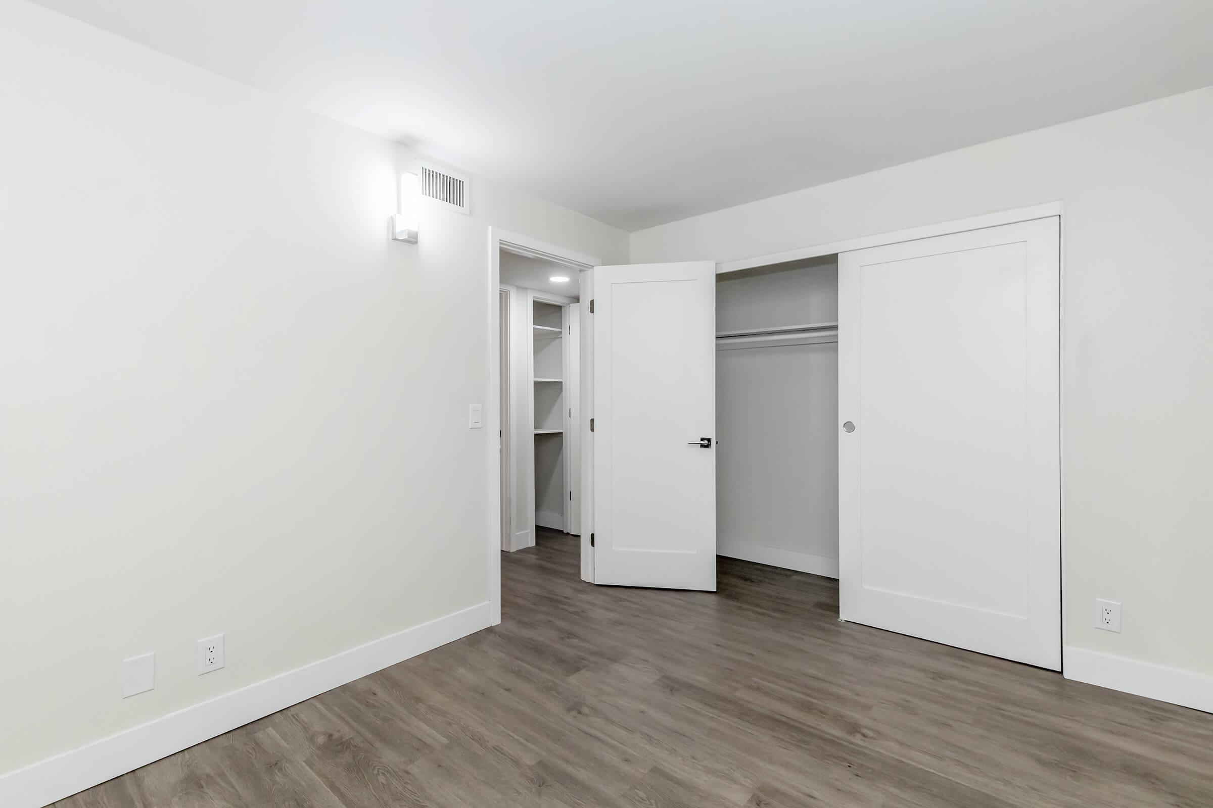 A well-lit, modern bedroom featuring light-colored walls, a wooden floor, and two closed doors leading to a closet. The room is empty with no furniture, showcasing a clean and spacious feel. A wall sconce provides additional lighting.