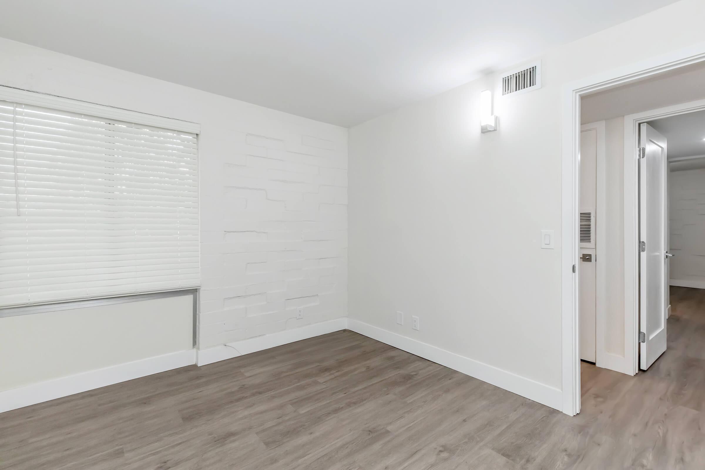 A bright, empty room with light-colored walls, a white textured accent wall, and large windows covered with white blinds. The flooring is light wood, and a wall-mounted light fixture is present. A doorway on the right leads to another area.