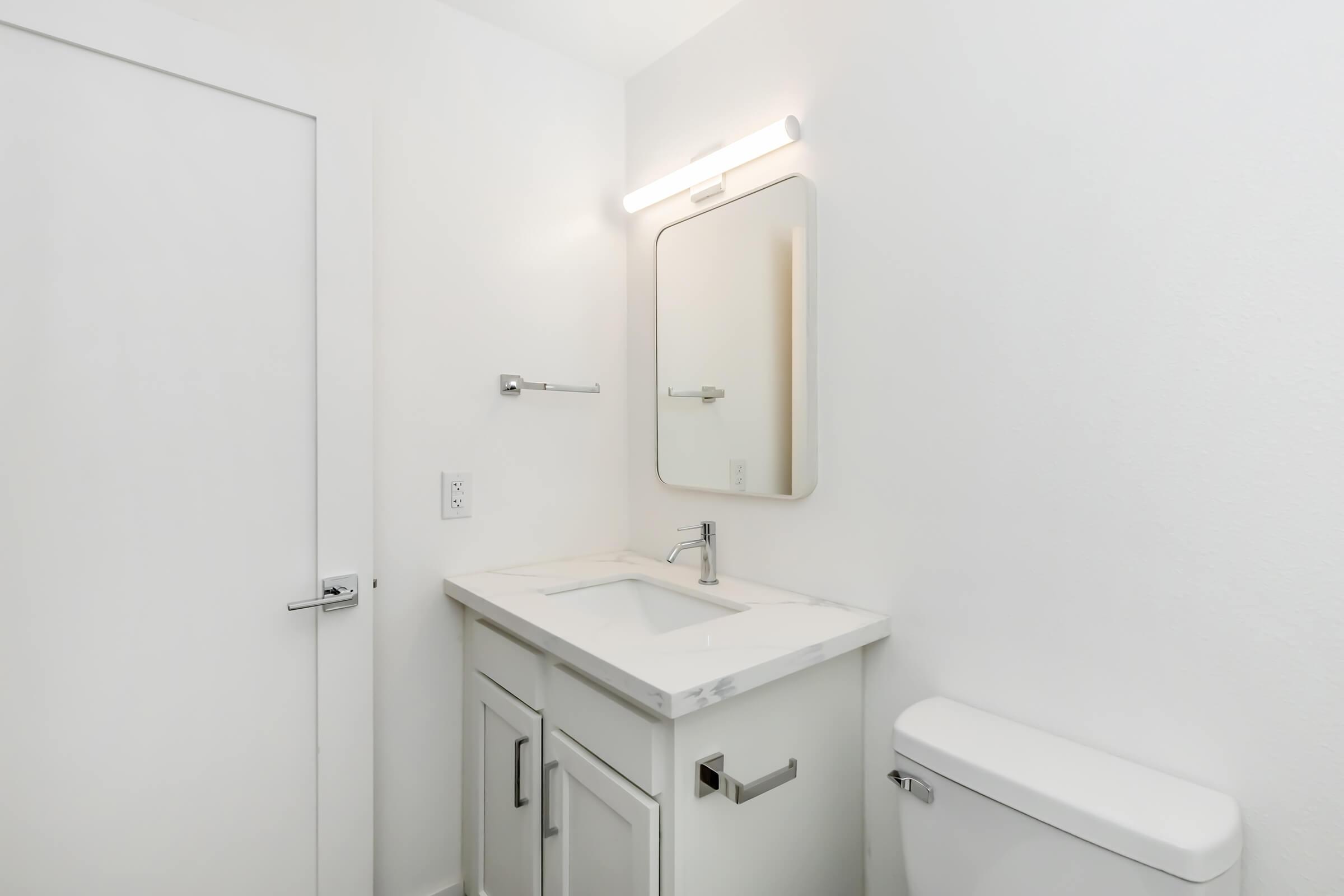 A clean and modern bathroom featuring a white vanity with a marble countertop, an oval mirror, a light fixture above, a wall-mounted towel bar, and a white toilet. The walls are painted white, and there is a white door visible in the background. The overall aesthetic is bright and minimalistic.