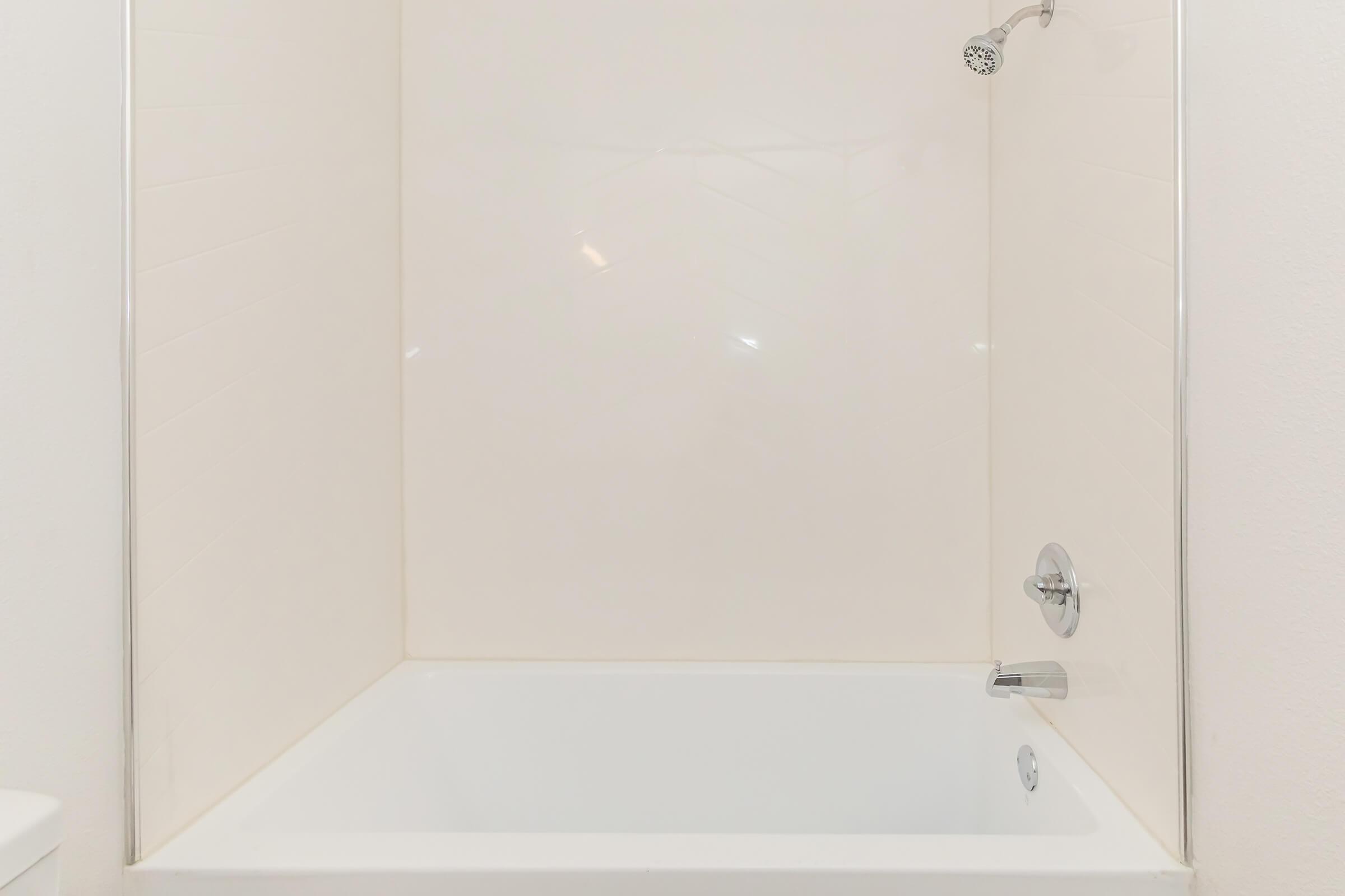 A clean, modern bathtub with a polished chrome showerhead mounted on a tiled wall. The space is well-lit, featuring a simple design with a plain white surface. The bathtub itself has a sleek, minimalist appearance with a single faucet.