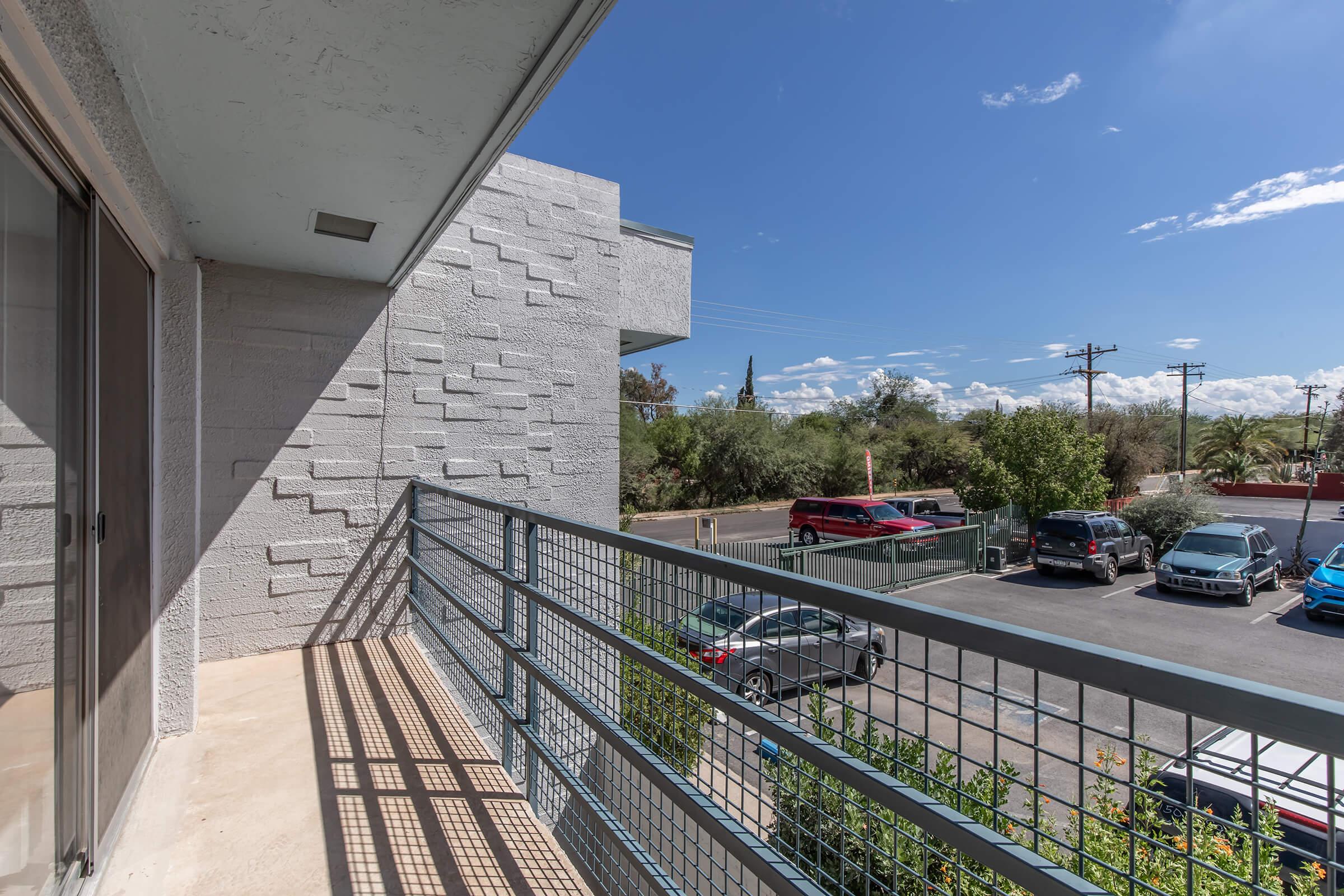A view from a balcony, featuring a metal railing overlooking a parking lot with several cars. In the background, there are sparse trees and blue skies with scattered clouds. The building's exterior has a textured wall.
