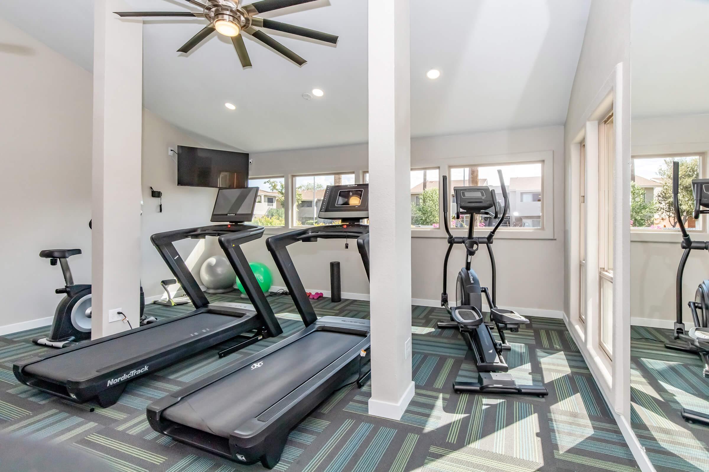 A modern fitness gym featuring two treadmills, an elliptical machine, and a stationary bike. Large windows provide natural light, and a ceiling fan circulates air. The room is decorated with a multi-colored patterned carpet and a green exercise ball is visible in the corner.