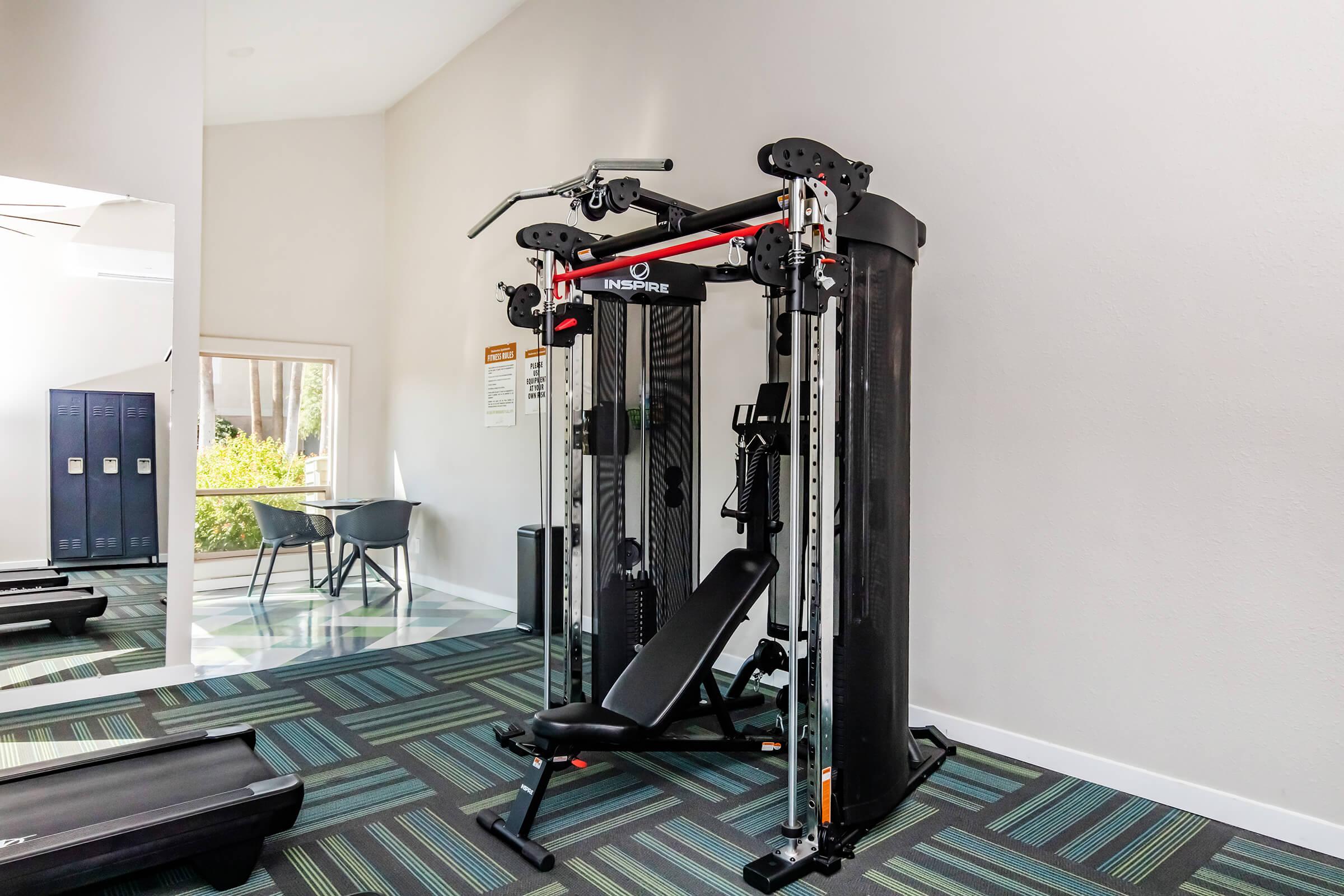 A modern gym space featuring a multi-station cable machine with adjustable settings, surrounded by a clean, bright environment. Visible are lockers in the background and a small seating area, creating a functional workout area.