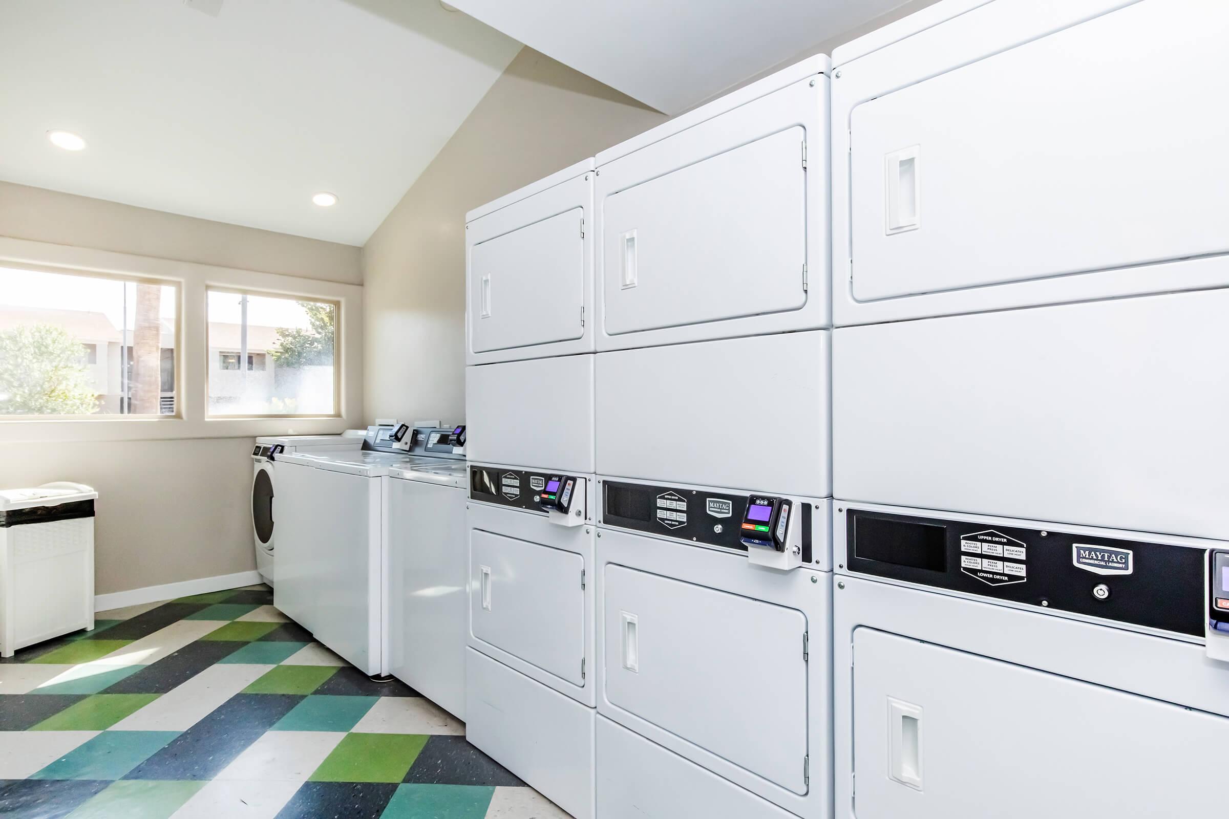 A bright and clean laundry room featuring white laundry machines, including washers and dryers. The room has large windows letting in natural light and is finished with a colorful, patterned floor. There are multiple white lockers for securing belongings, creating an organized space for laundry activities.