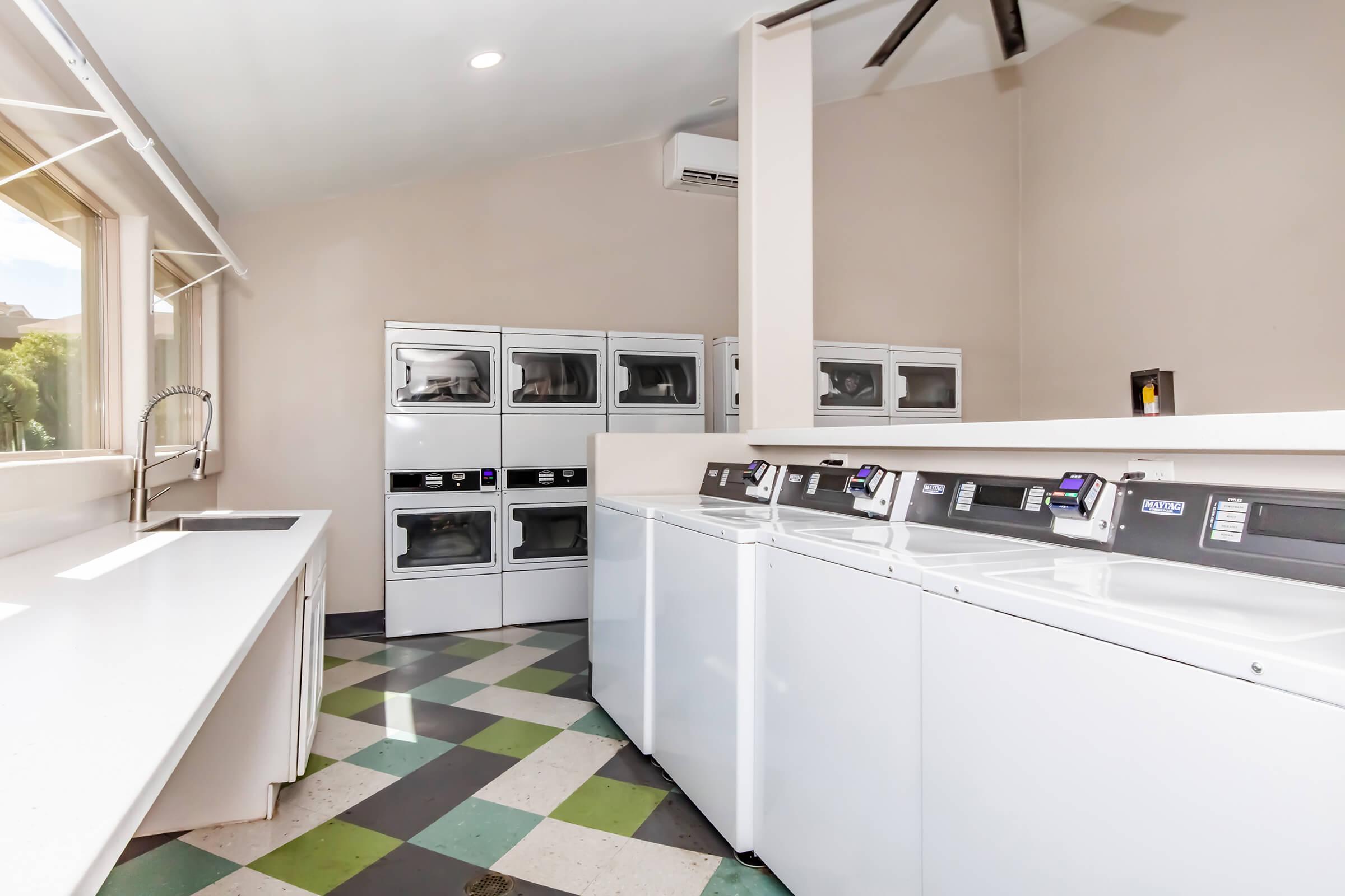 A modern laundry room featuring multiple white washers and dryers, a countertop workspace, and large windows allowing natural light. The floor is patterned with black and green tiles, and there is a ceiling fan installed. The space is clean and organized, designed for efficiency.