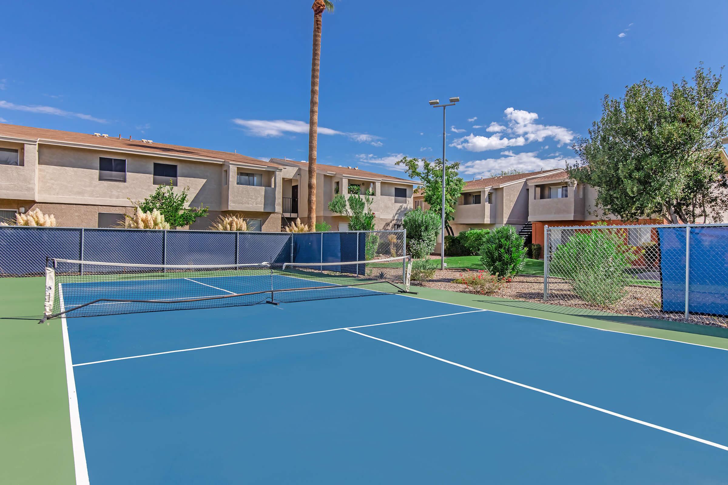 A tennis court with a blue surface, surrounded by a fence, and backed by residential buildings. Lush greenery and trees are visible in the background, under a clear blue sky with a few clouds. The court is empty, ready for play.