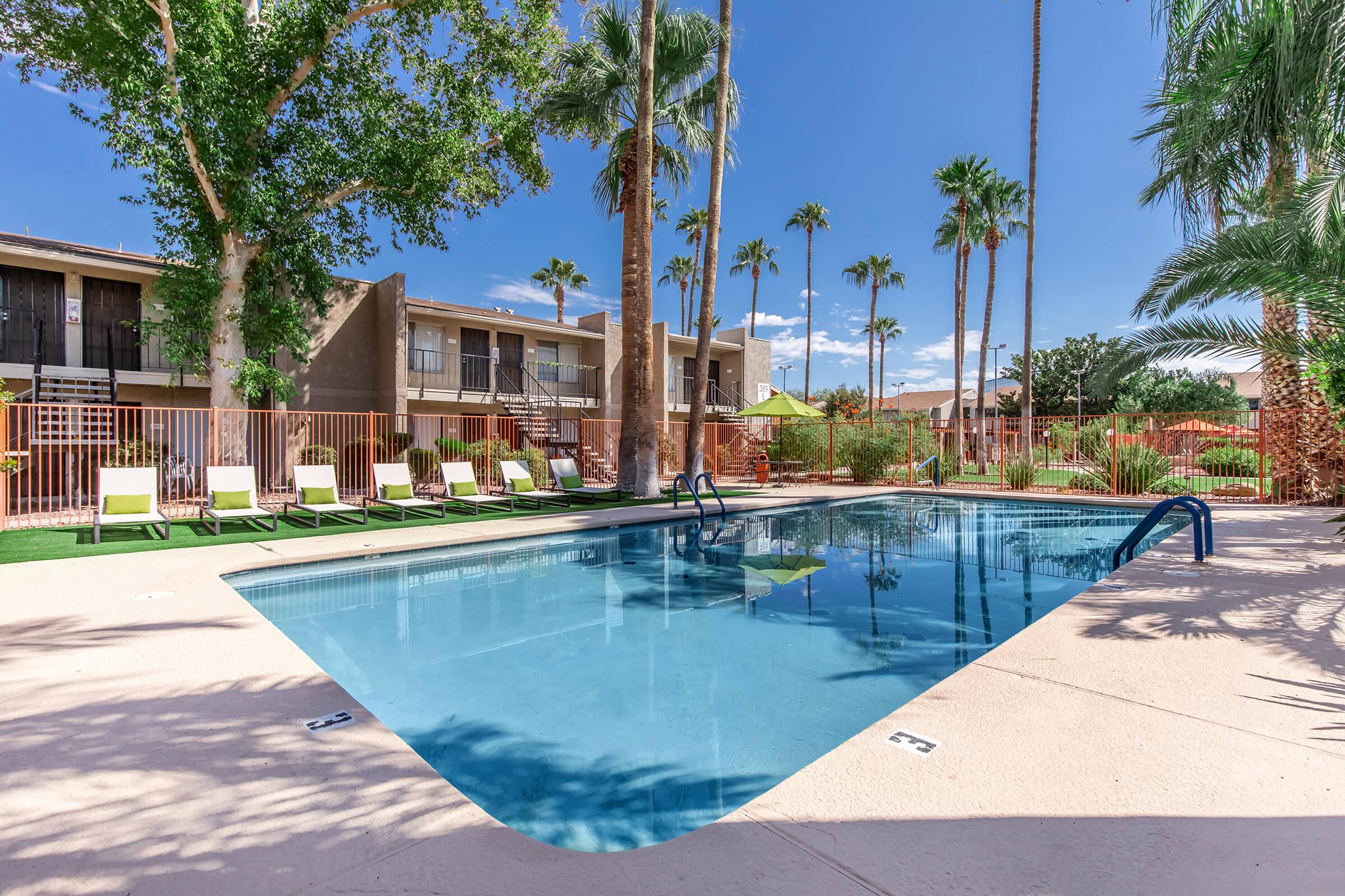 A clear swimming pool surrounded by palm trees and lounge chairs, with a backdrop of two-story buildings under a bright blue sky. The pool area features green grass and a fence separating it from the surrounding area, creating a relaxing outdoor space.
