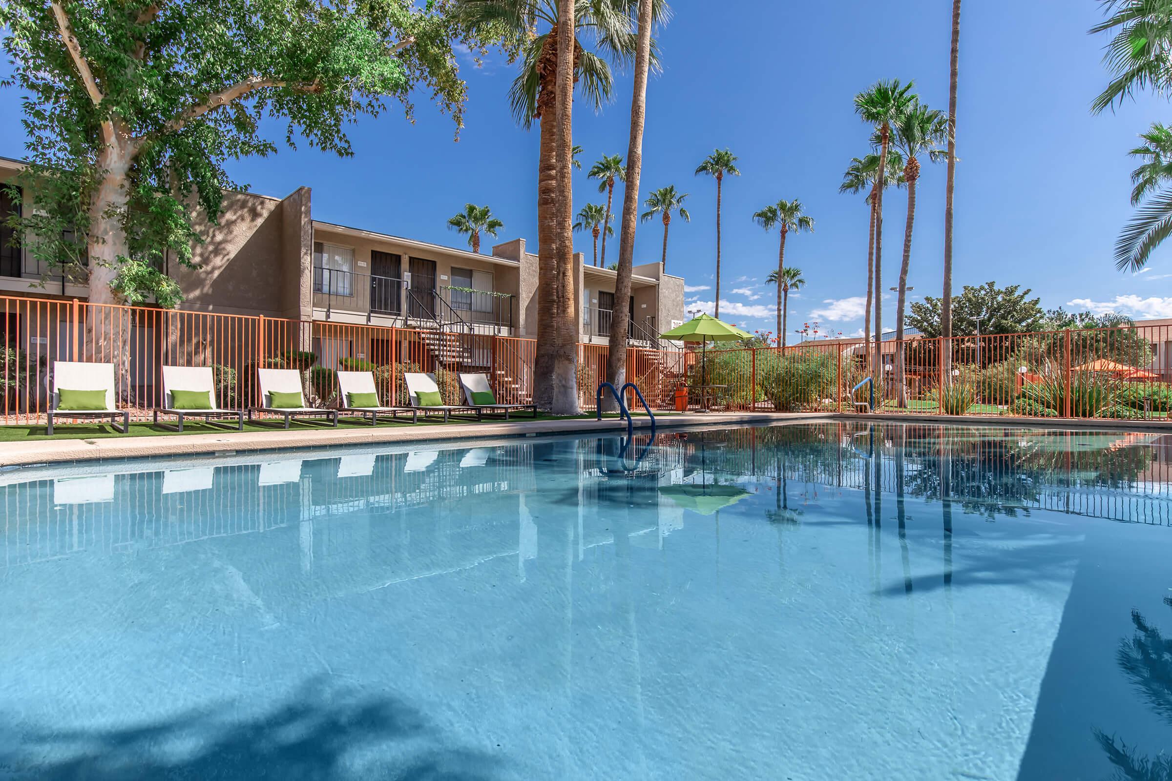 A clear blue swimming pool surrounded by palm trees and lounge chairs. In the background, there are two-story buildings with balconies under a bright blue sky. The scene conveys a relaxing outdoor atmosphere, perfect for leisure and enjoyment.