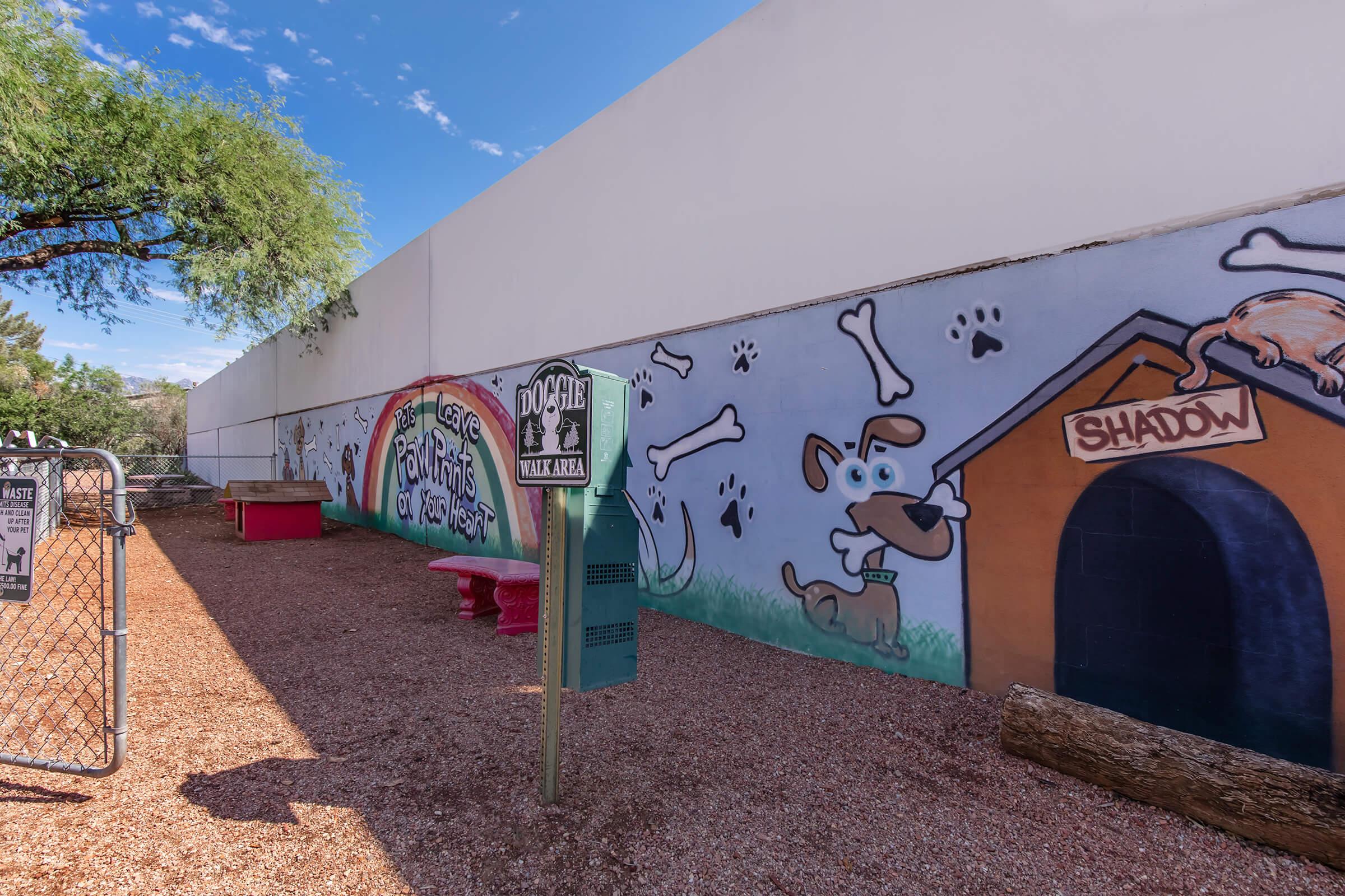 A dog park area featuring colorful murals of dogs, bones, and a doghouse on the wall. The space includes red benches, a sign marking the dog park, and a gravel ground with trees nearby. The atmosphere is playful and inviting for pets and their owners.