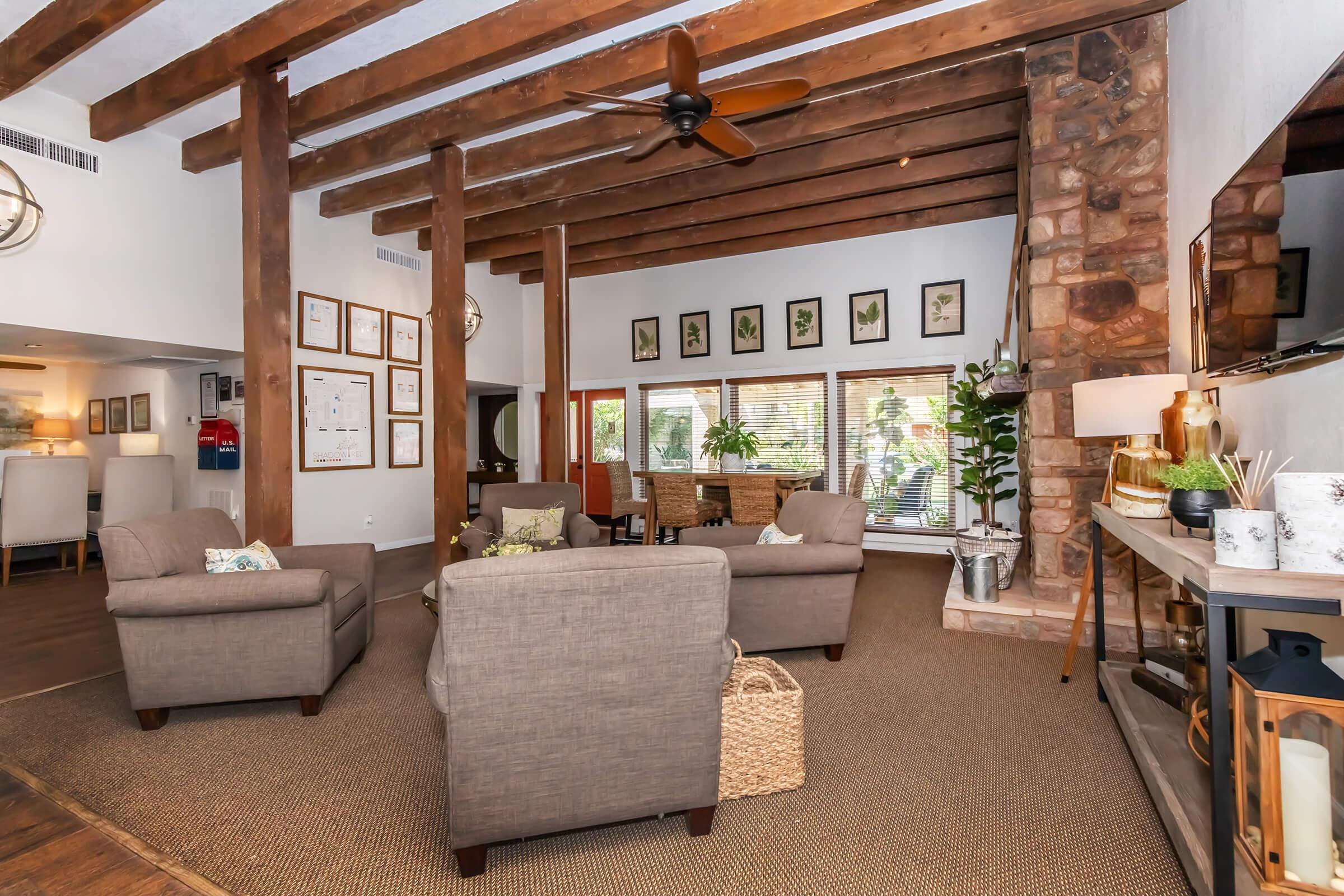 Cozy interior of a living space featuring wooden beams, a stone fireplace, and comfortable gray armchairs. The room is decorated with plants and framed artwork on the walls, creating a warm and inviting atmosphere. Natural light spills in through large windows, enhancing the welcoming vibe.