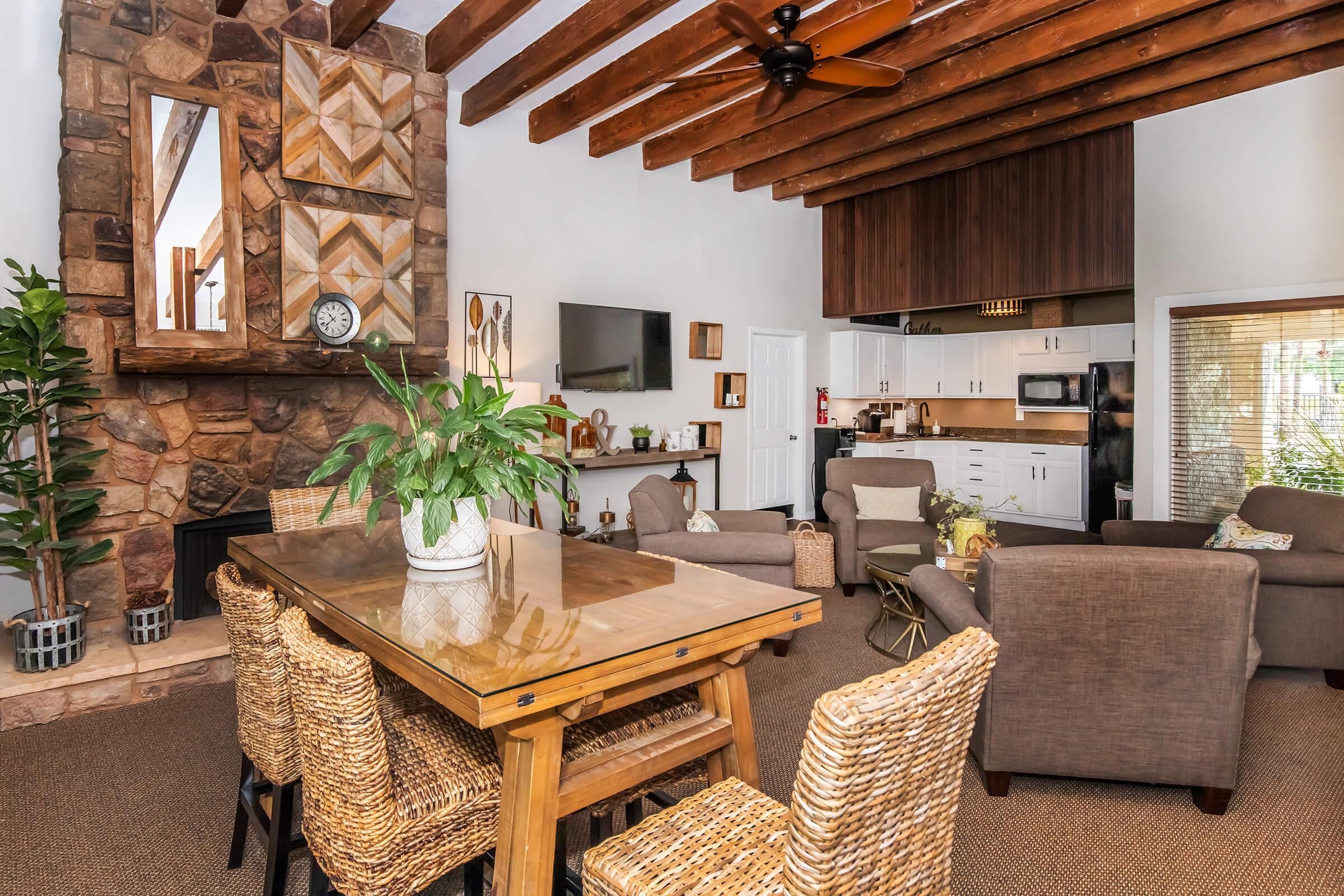 A cozy living space featuring wooden beams and stone accents. It includes a dining table surrounded by woven chairs, a comfortable seating area with armchairs and a sofa, a wall-mounted TV, and a few decorative plants. The overall atmosphere is warm and inviting with natural light coming in.