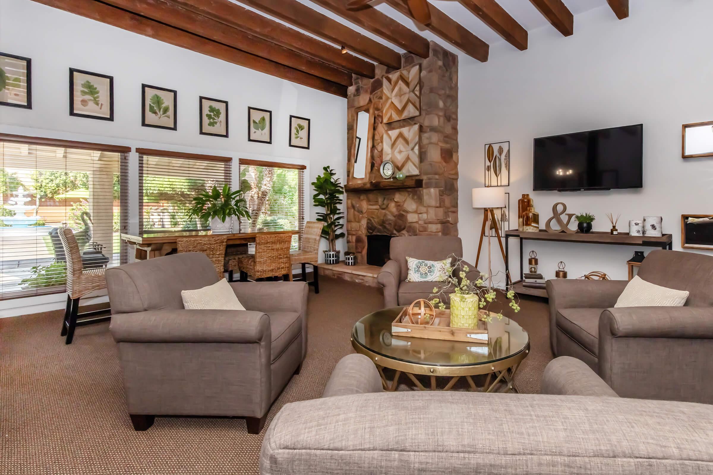 A cozy living room featuring two armchairs and a couch, with a stone fireplace as the focal point. The walls are adorned with framed botanical art, and large windows offer natural light. A wooden dining table and decorative plants are visible in the background, creating a warm and inviting atmosphere.