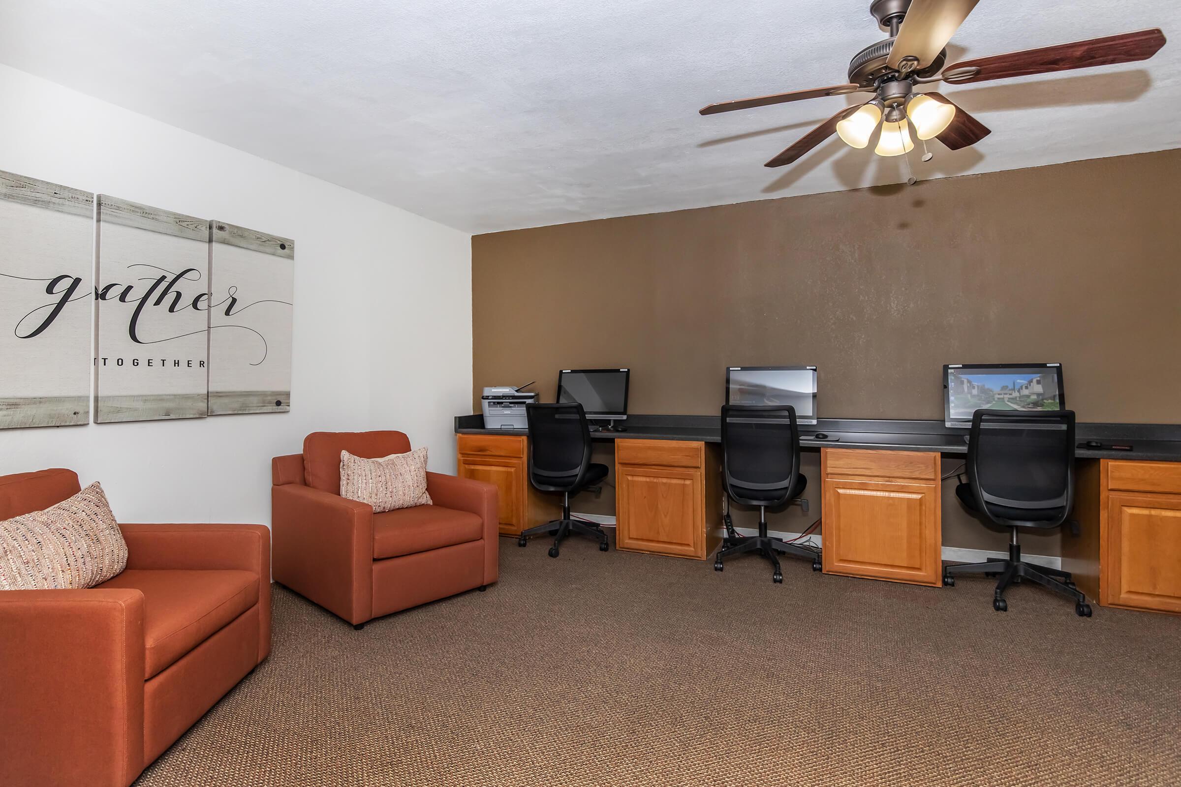 A cozy office space featuring two computer stations with black chairs against a brown wall. There's a decorative "gather together" sign on a wooden panel, and a brown armchair with a patterned cushion is placed nearby. The room has soft lighting and carpeted flooring, creating a welcoming atmosphere.