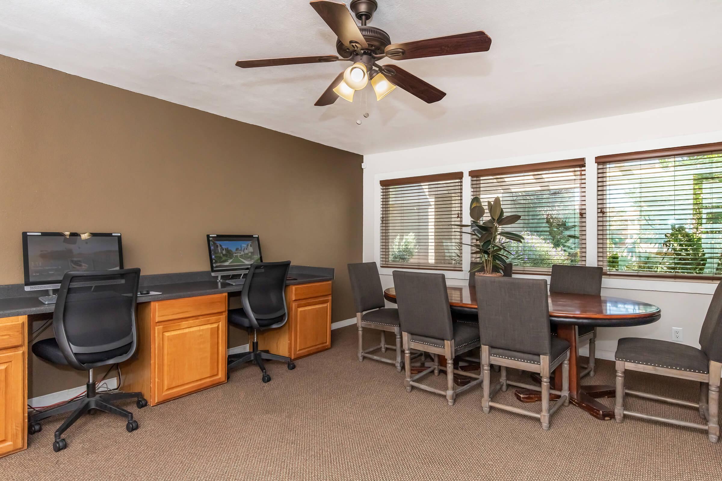 A modern office space featuring two computer desks with monitors, ergonomic chairs, and a round dining table surrounded by gray upholstered chairs. The room is well-lit with natural light coming through large windows and has a ceiling fan for ventilation. The walls are painted in a warm, neutral tone.