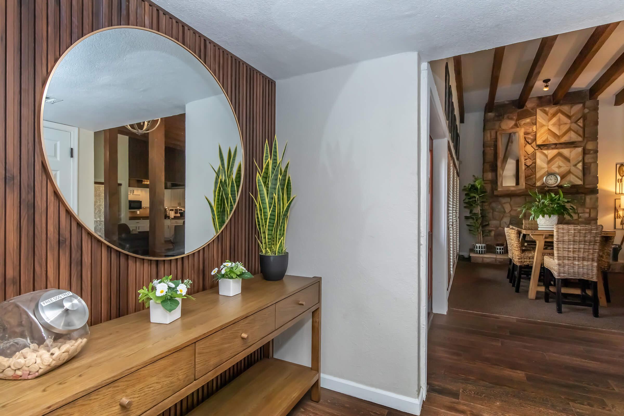 A modern hallway featuring a circular mirror above a wooden console table. The table displays small potted plants and a glass jar of corks. On one side, there is a tall green plant, and the wall has warm wood paneling. In the background, an open space leads to a cozy living area with stone accents.