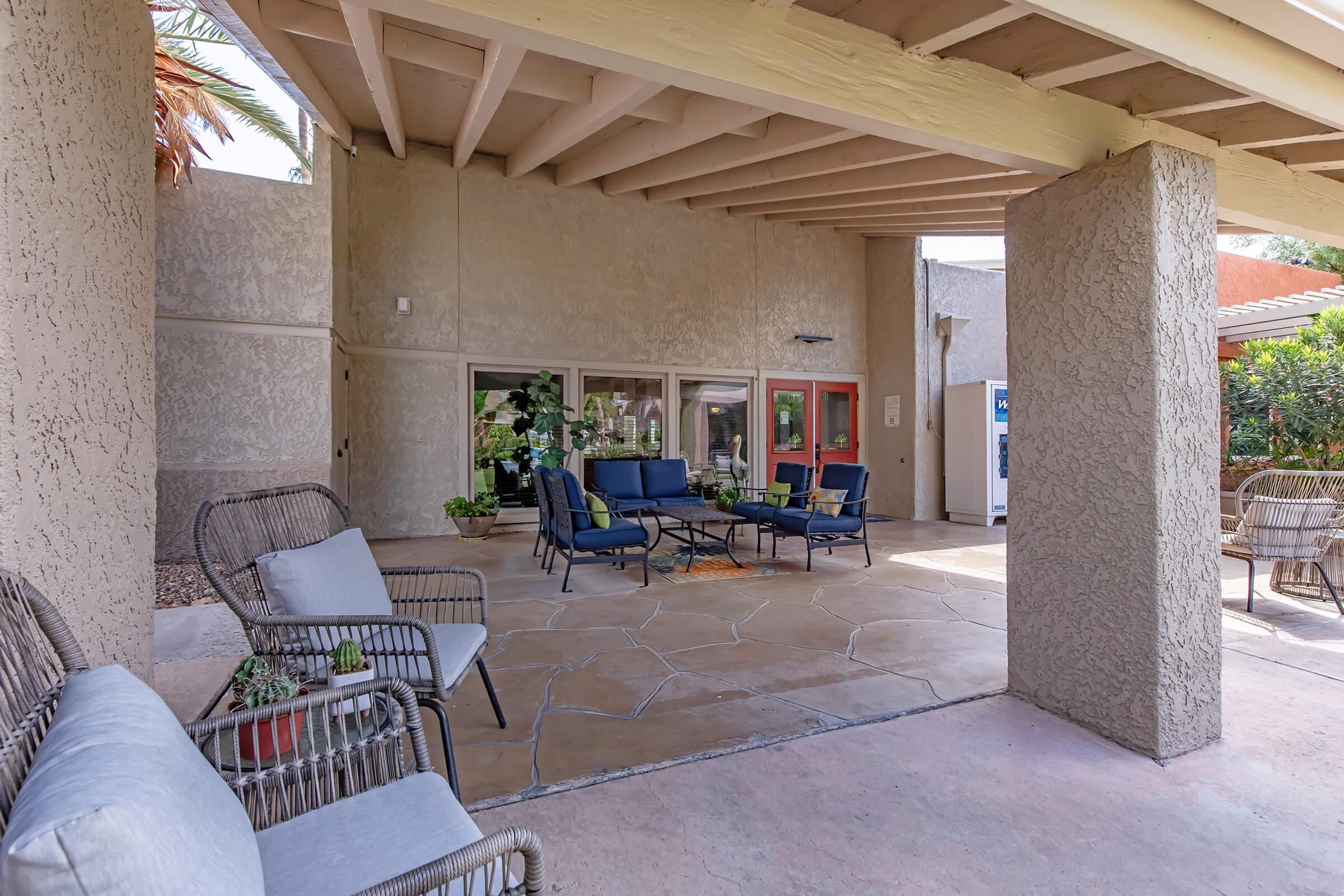A cozy outdoor patio featuring wicker chairs and a small table, surrounded by greenery. The area is sheltered by a roof, with a view of large windows and doors to an indoor space. Sunlight streams in, creating a warm and inviting atmosphere for relaxation.